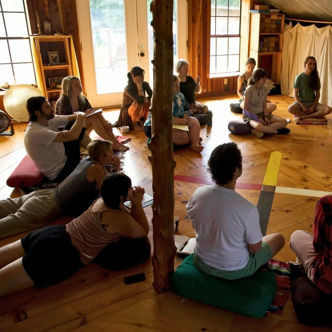 People sitting on the floor and cushions in a circle, participating in a group meditation or yoga session in a cozy wooden room with large windows.