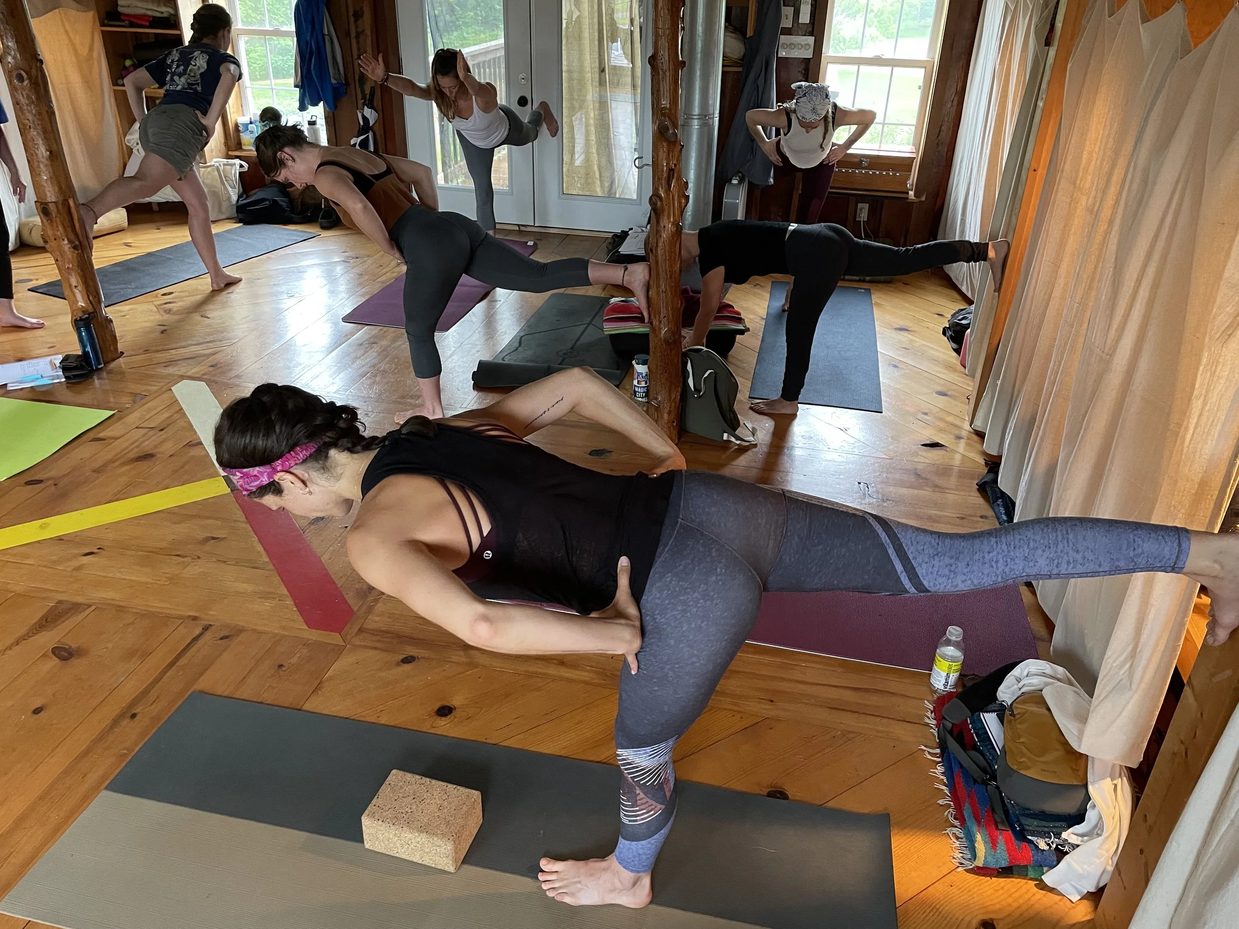 Group of women practicing yoga in a cozy wooden room with large windows showing greenery outside. They are in various yoga poses on mats, with some using blocks for support.