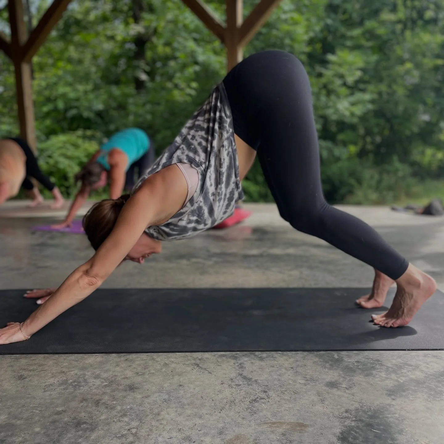 A woman practicing yoga outdoors on a black mat, in a downward dog position during a group yoga class, with other participants in similar poses, under a wooden pavilion surrounded by green trees.