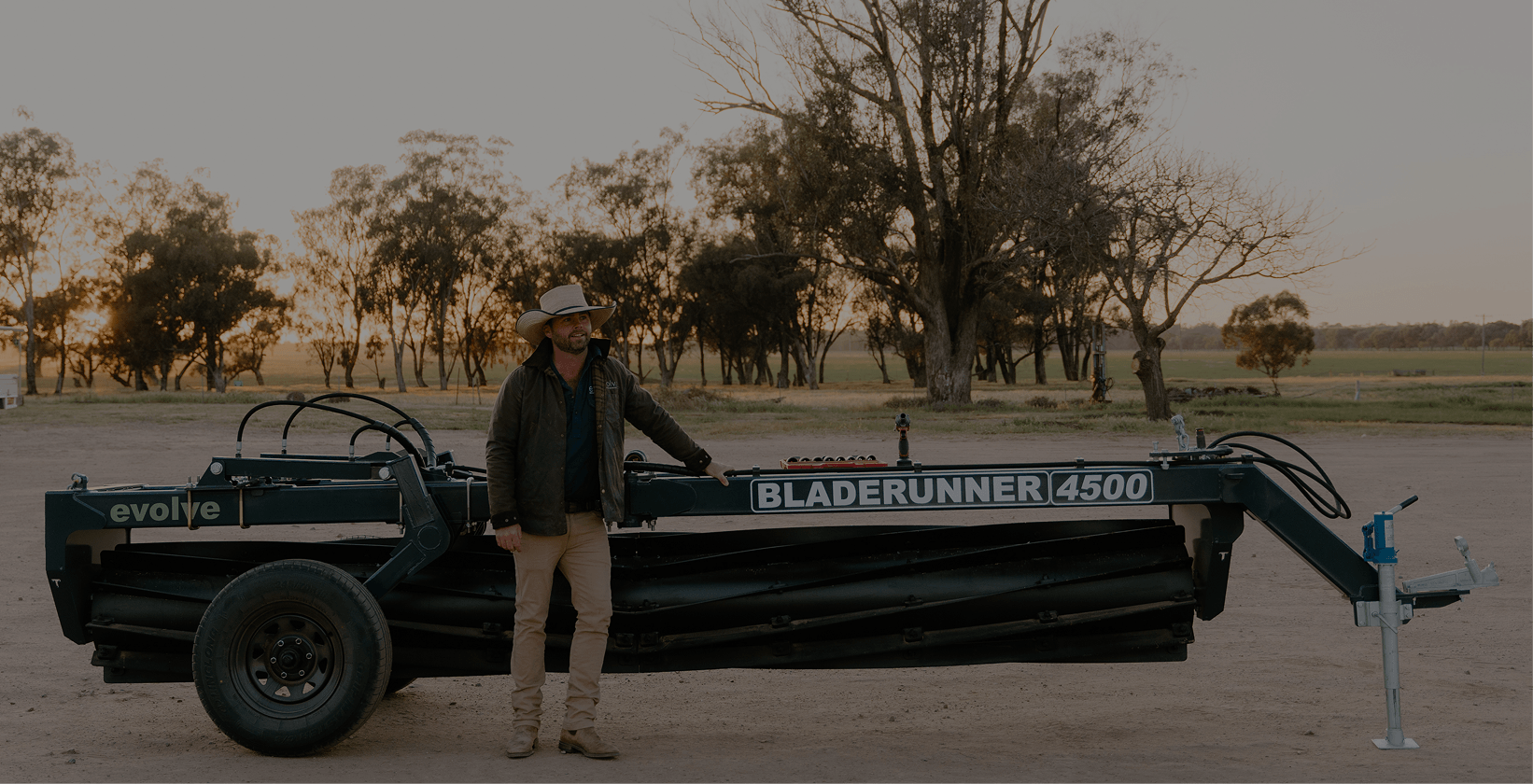 A man standing next to a large farm implement labeled 'Bladerunner 4500' on a dirt field with trees in the background during sunset.