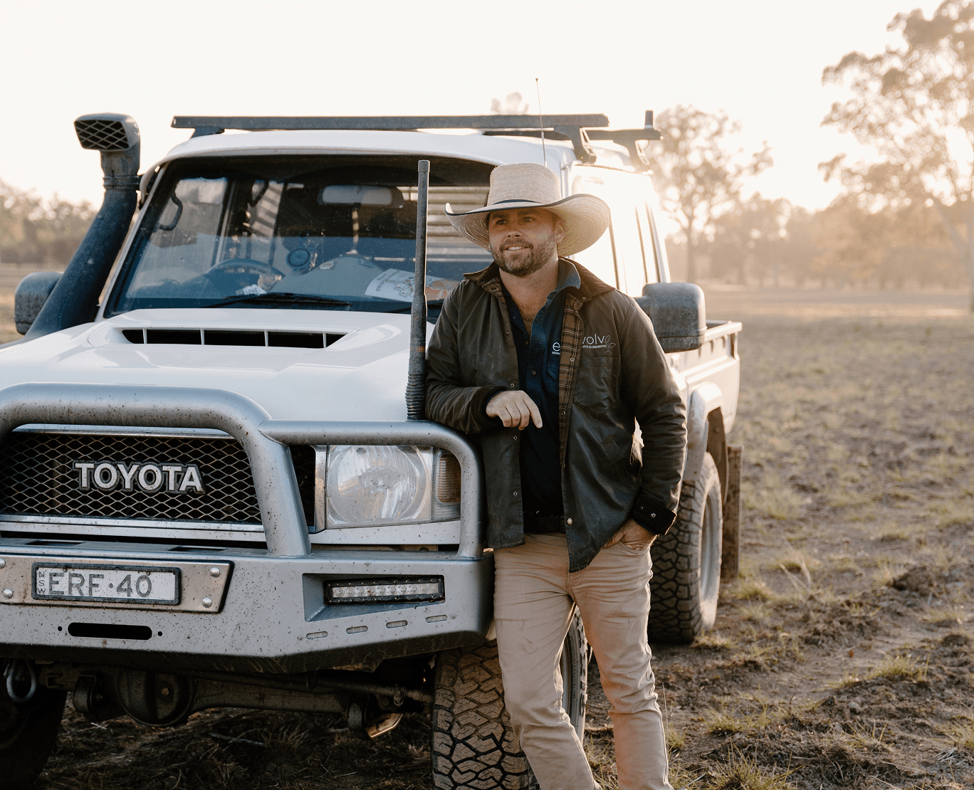 A man wearing a cowboy hat and casual outdoor clothing leaning on the front of a white Toyota pickup truck in a rural field during sunset.