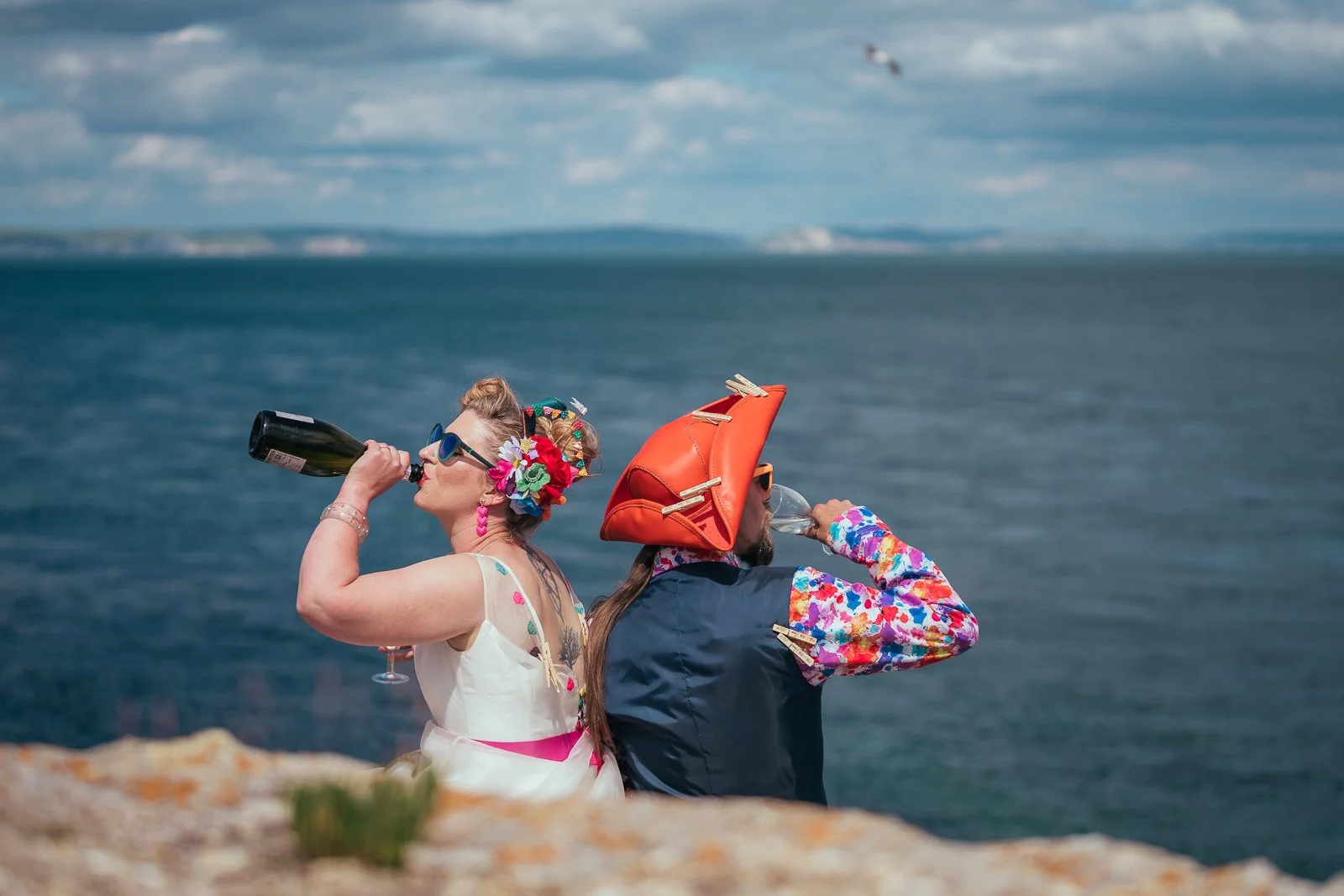 Two people in colorful costumes and headwear drinking from bottles by the ocean on a cloudy day.