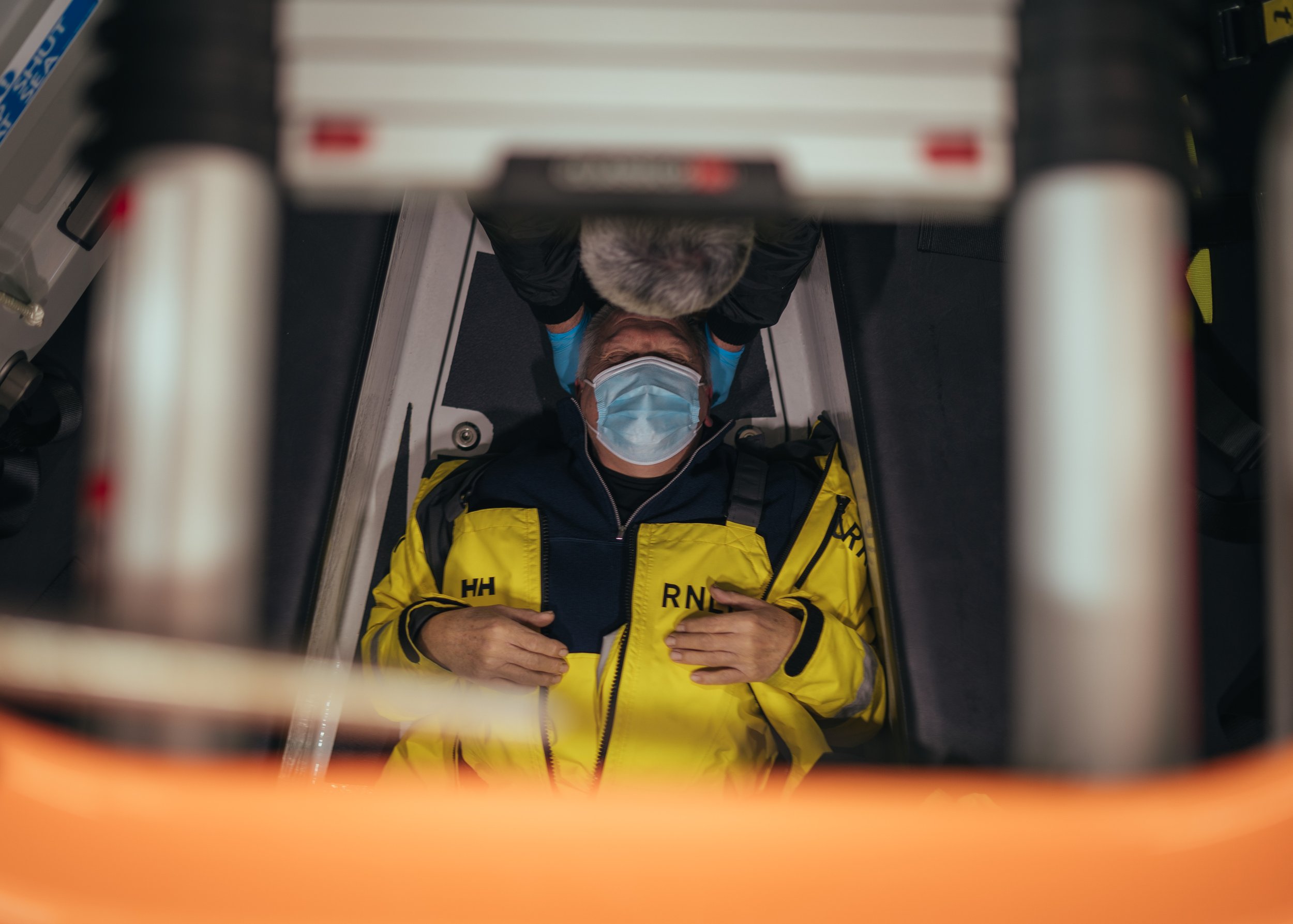 Emergency medical technician lying on a gurney, wearing a yellow jacket with 'RNC' and 'HH' on it, mask, and gloves, being attended to by another person.