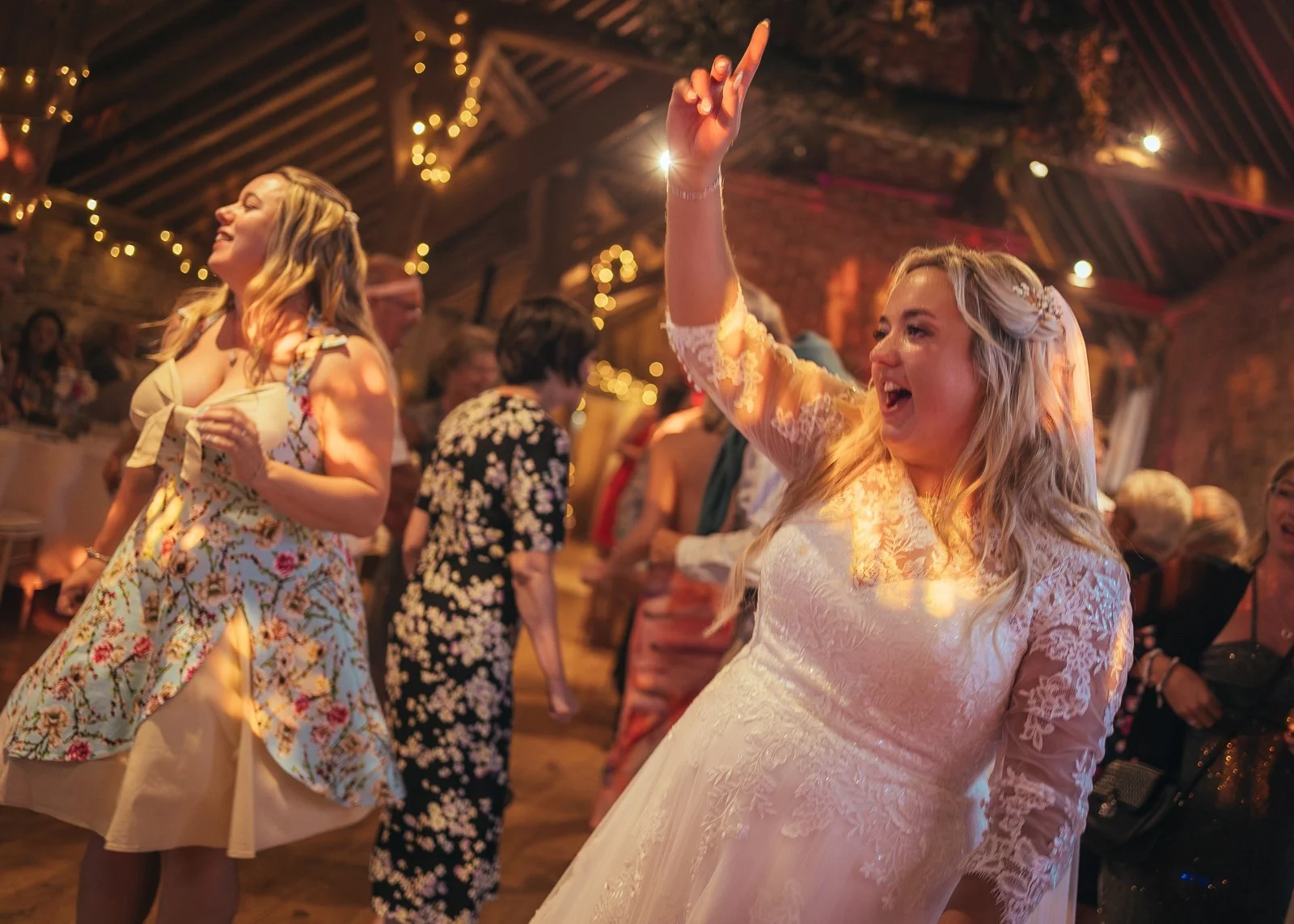 A bride in a lacy white wedding dress is dancing and smiling, with her hand raised, in a warmly lit venue with string lights and friends dancing in the background.