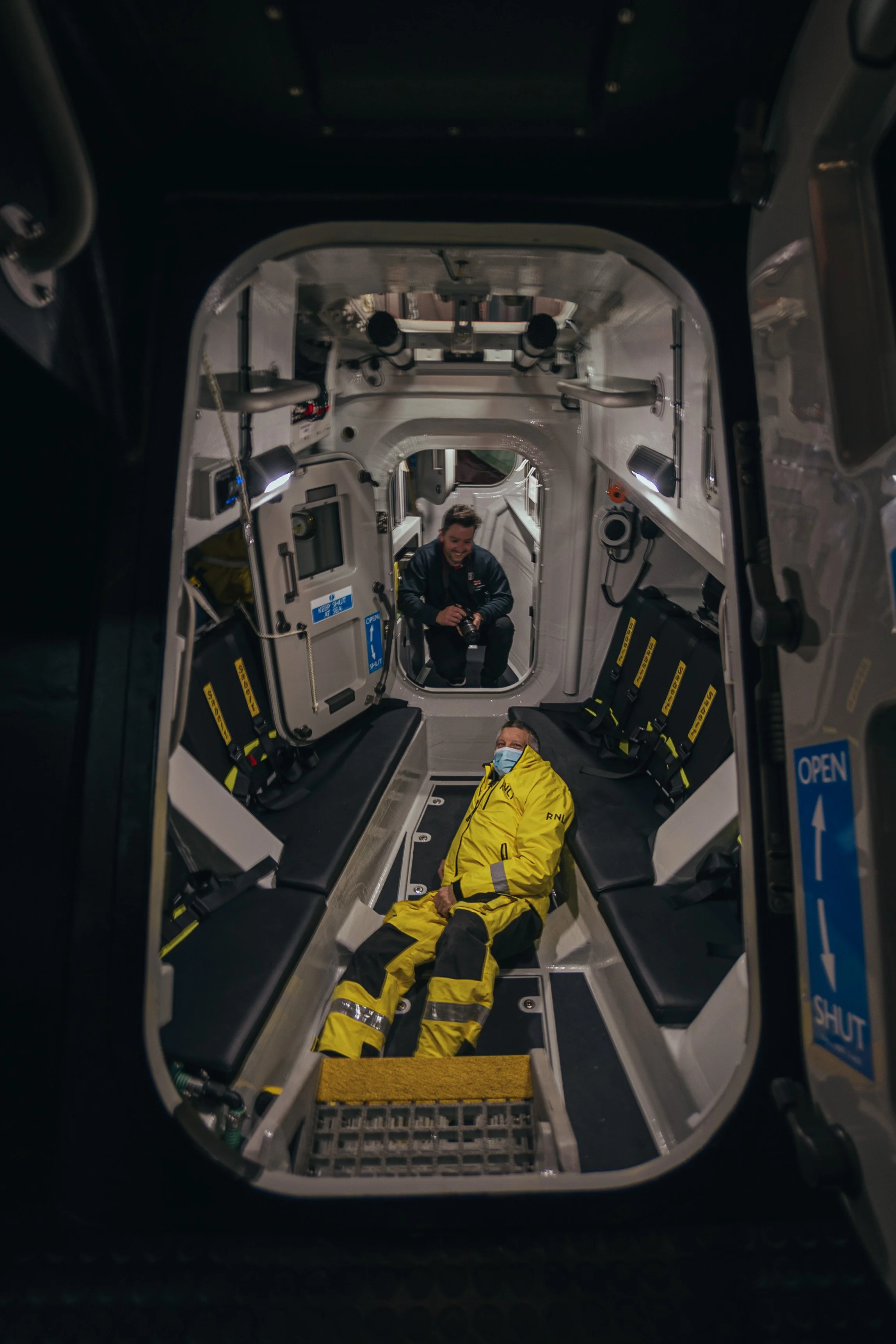 Inside an ambulance, a paramedic in a yellow uniform is lying on the floor wearing a face mask, while another person with a camera is sitting in the background near the doorway, smiling.
