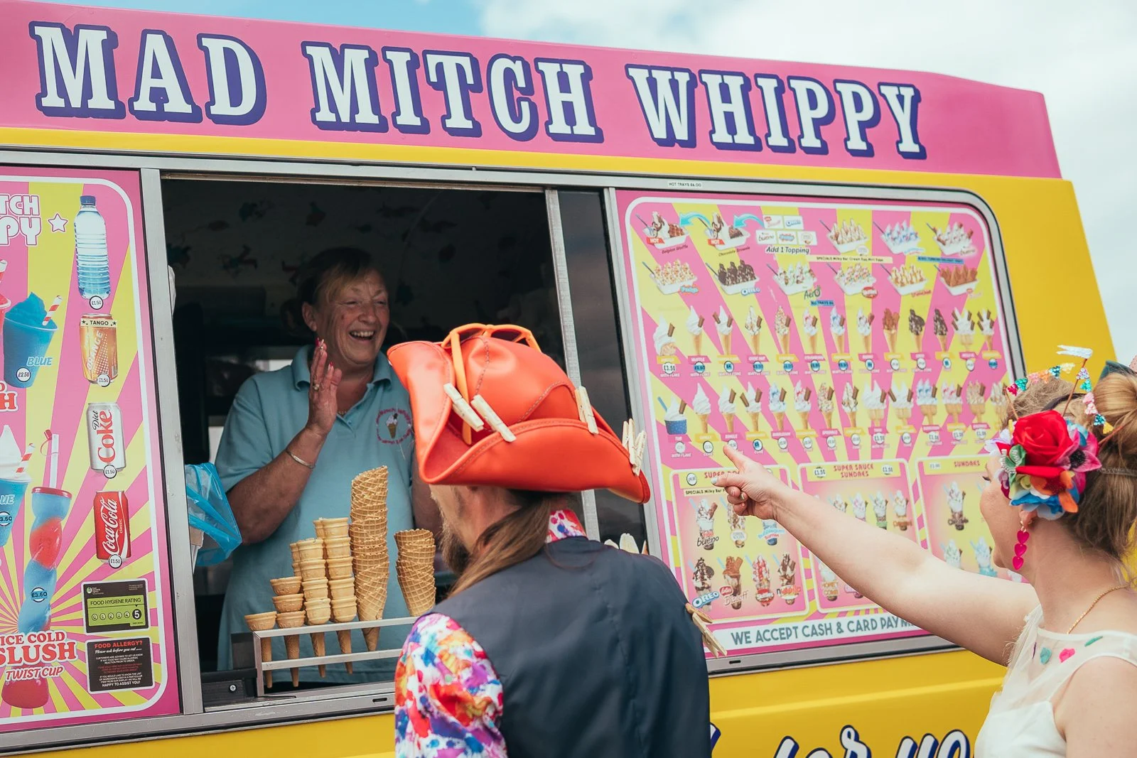 A woman laughing and talking to customers at an ice cream truck named 'Mad Mitch Whippy'. The truck has a colorful menu and several ice cream cones on display. Groom and bride points at the menu, wearing a red flower headband and floral earrings.