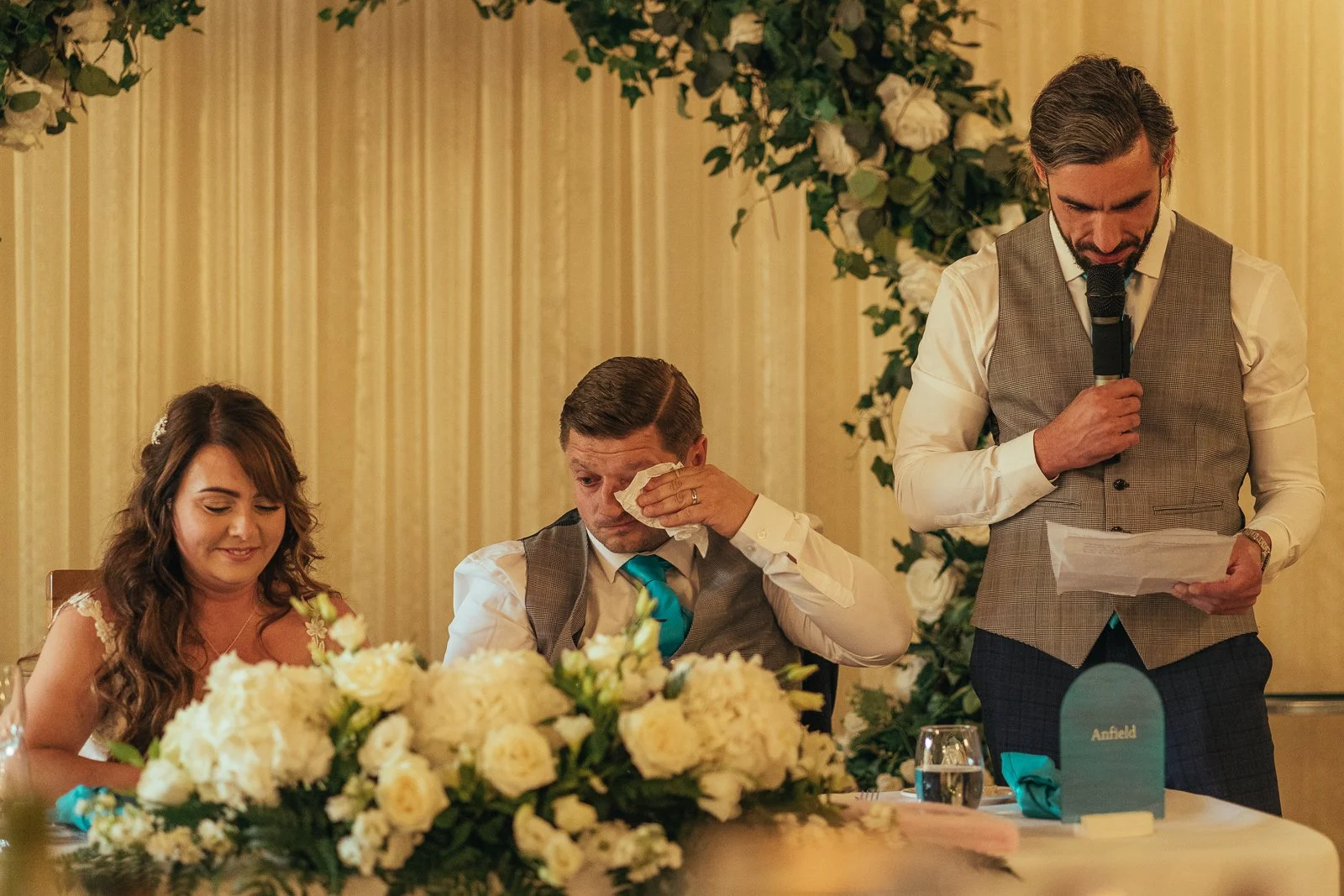 A bride, groom, and best man at a wedding reception. The bride is smiling, the groom is wiping tears with a tissue, and the best man is reading from a paper while holding a microphone. The table has a large floral arrangement.