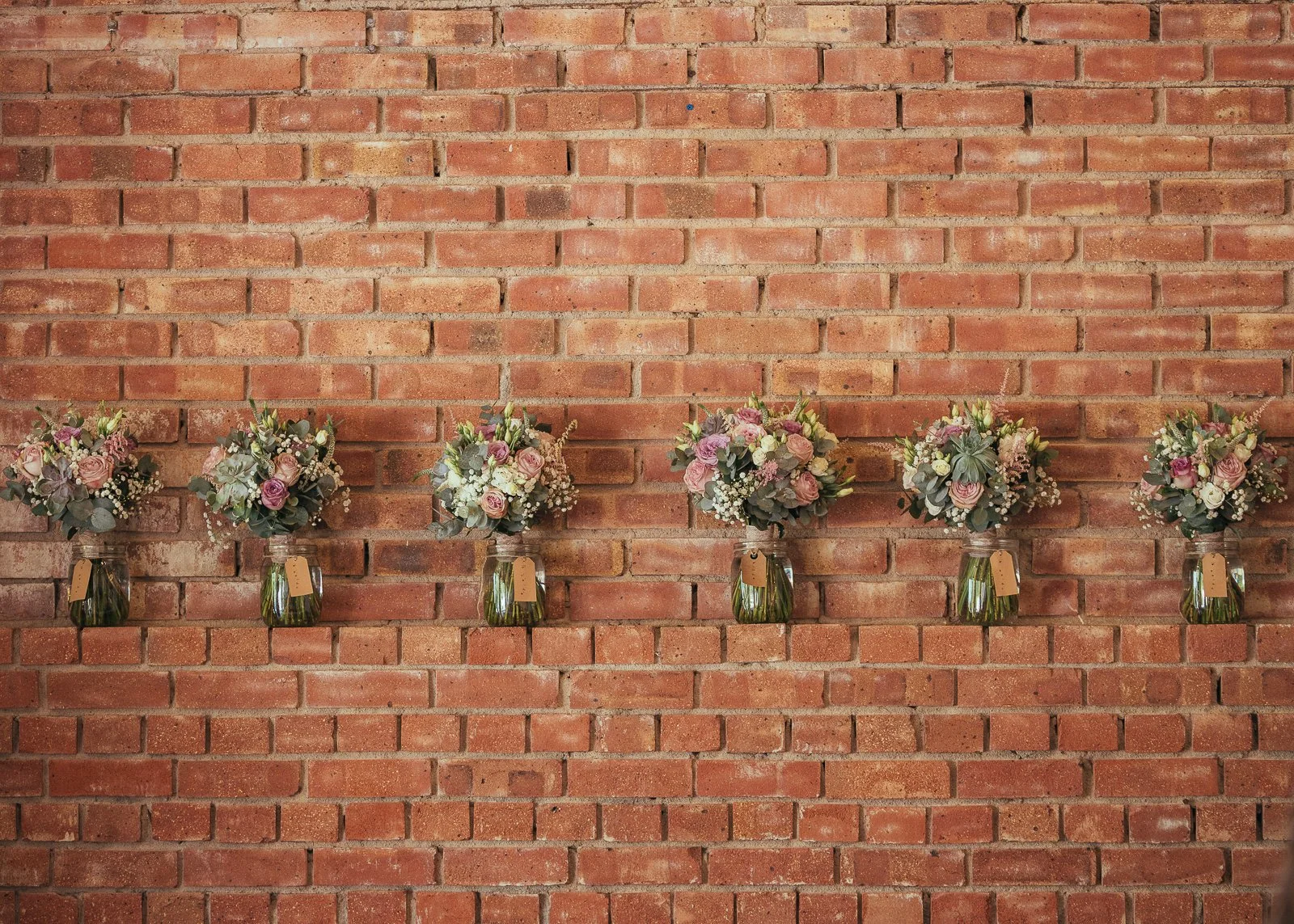 Six flower arrangements in glass jars mounted on a brick wall, with pink, white, and purple flowers including roses and baby's breath.