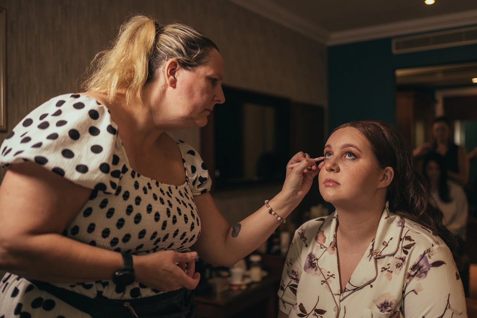 Woman applying makeup to another woman in a room, with a background that includes a person taking a photo.