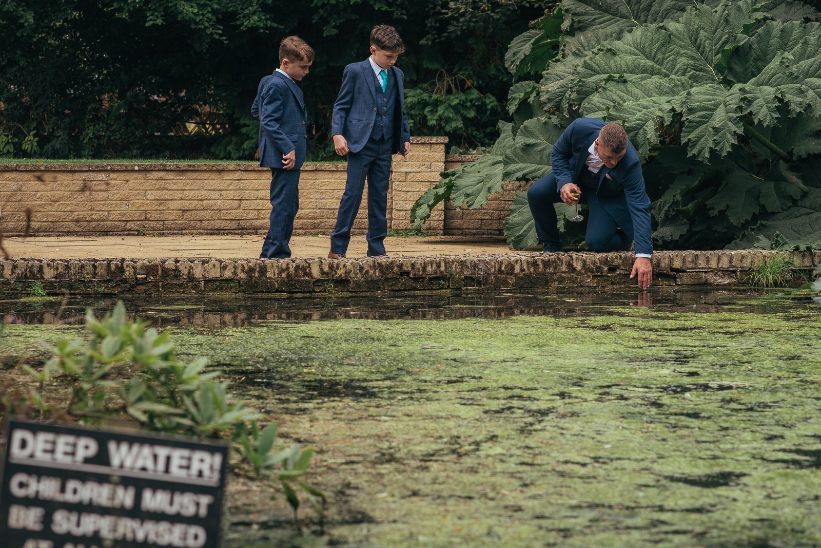 A man in a suit kneeling by a pond with two young boys in suits watching him examine something in the water, with a sign in the foreground indicating deep water children must be supervised.