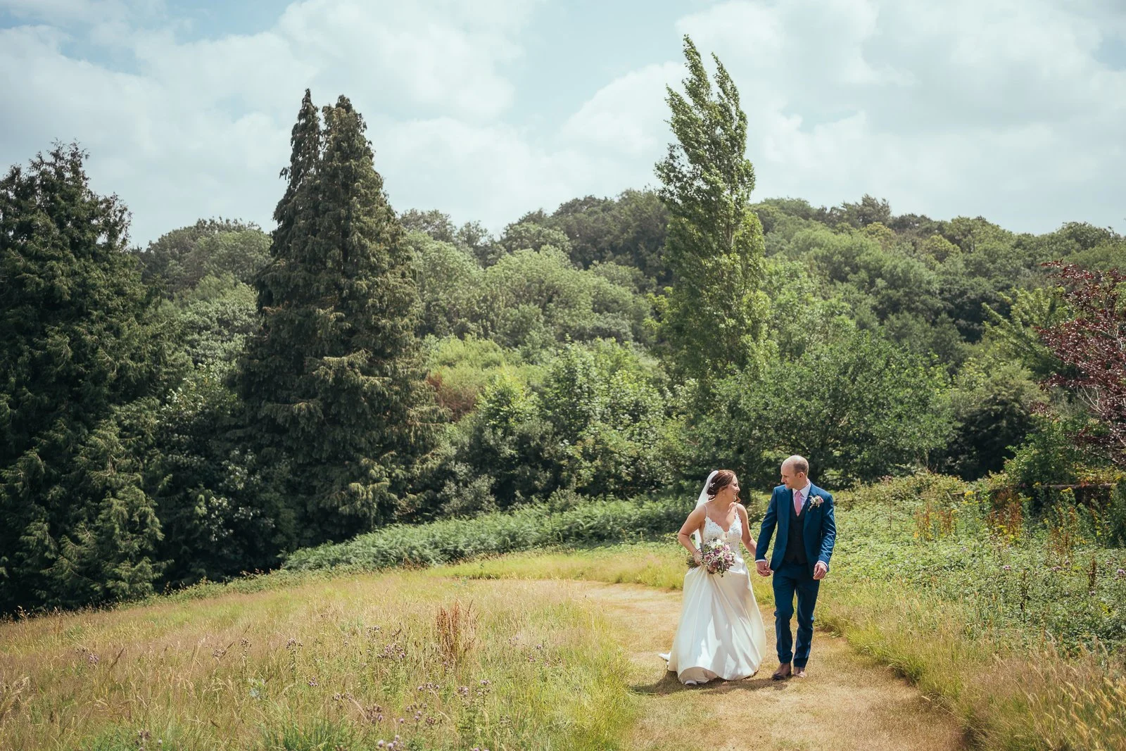 Bride and groom walking hand in hand on a grassy trail surrounded by green trees and foliage during their wedding day.
