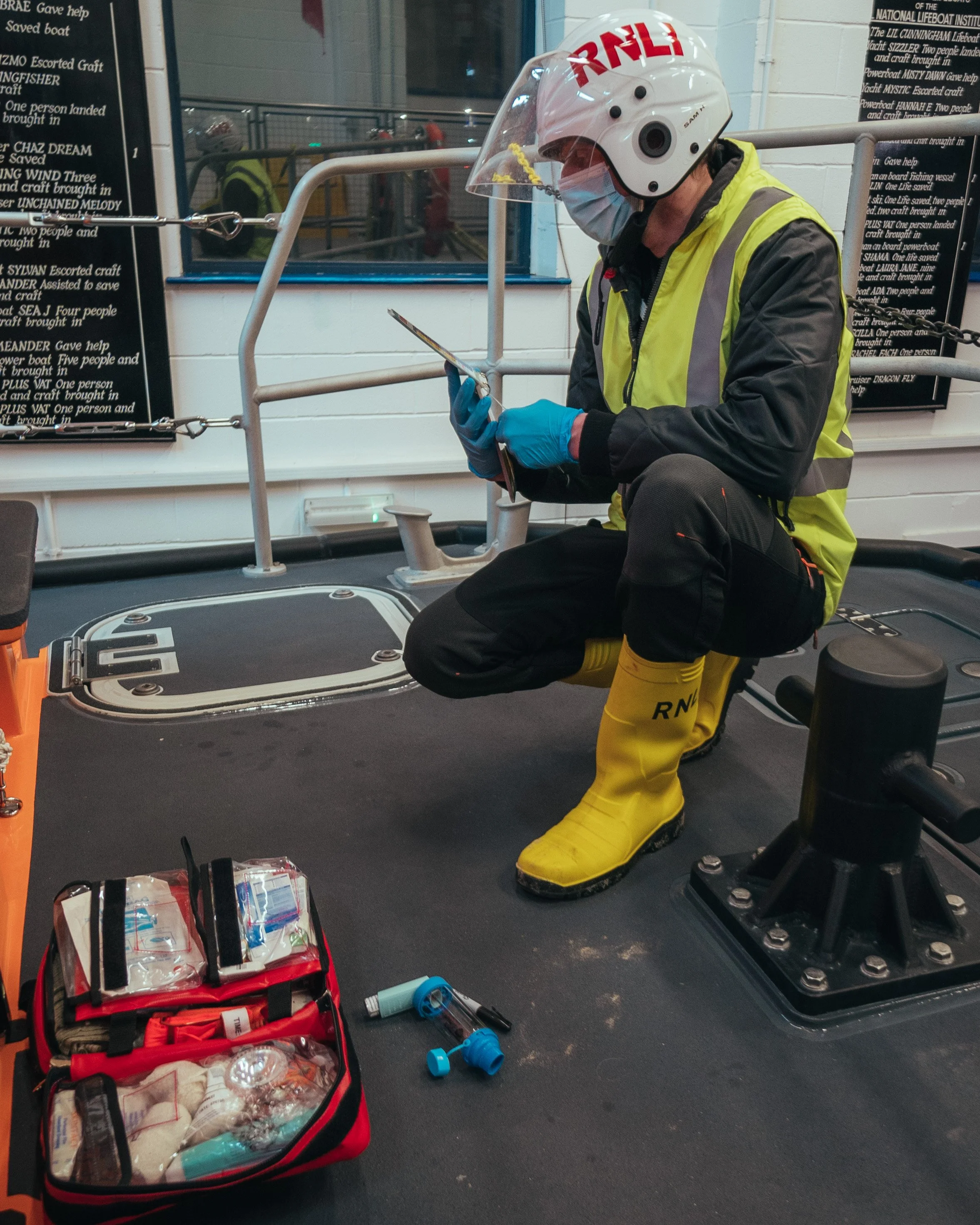 A person dressed in safety gear including a helmet, face mask, gloves, and yellow rubber boots, is crouching on a boat deck and looking at a tablet. In the background, there are safety signs, equipment, and a window with reflections.
