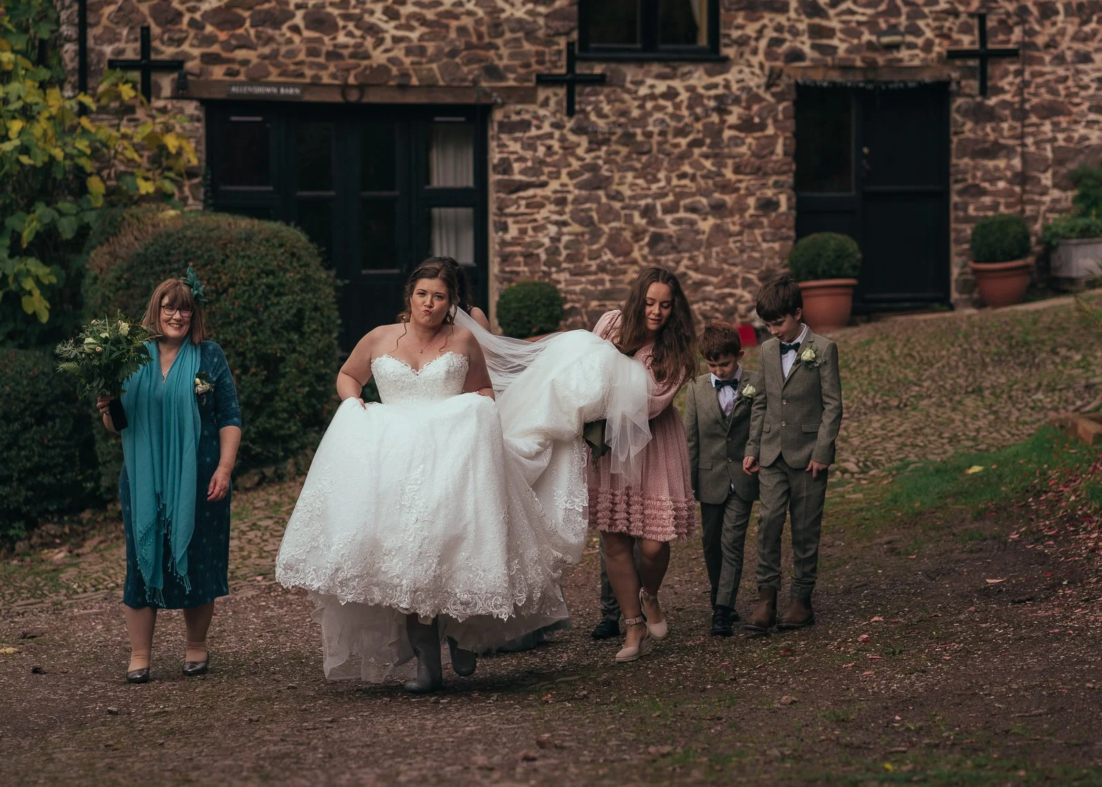 Group of people walking outdoors, woman in wedding dress in the center, others assisting, in front of a stone building with potted plants.