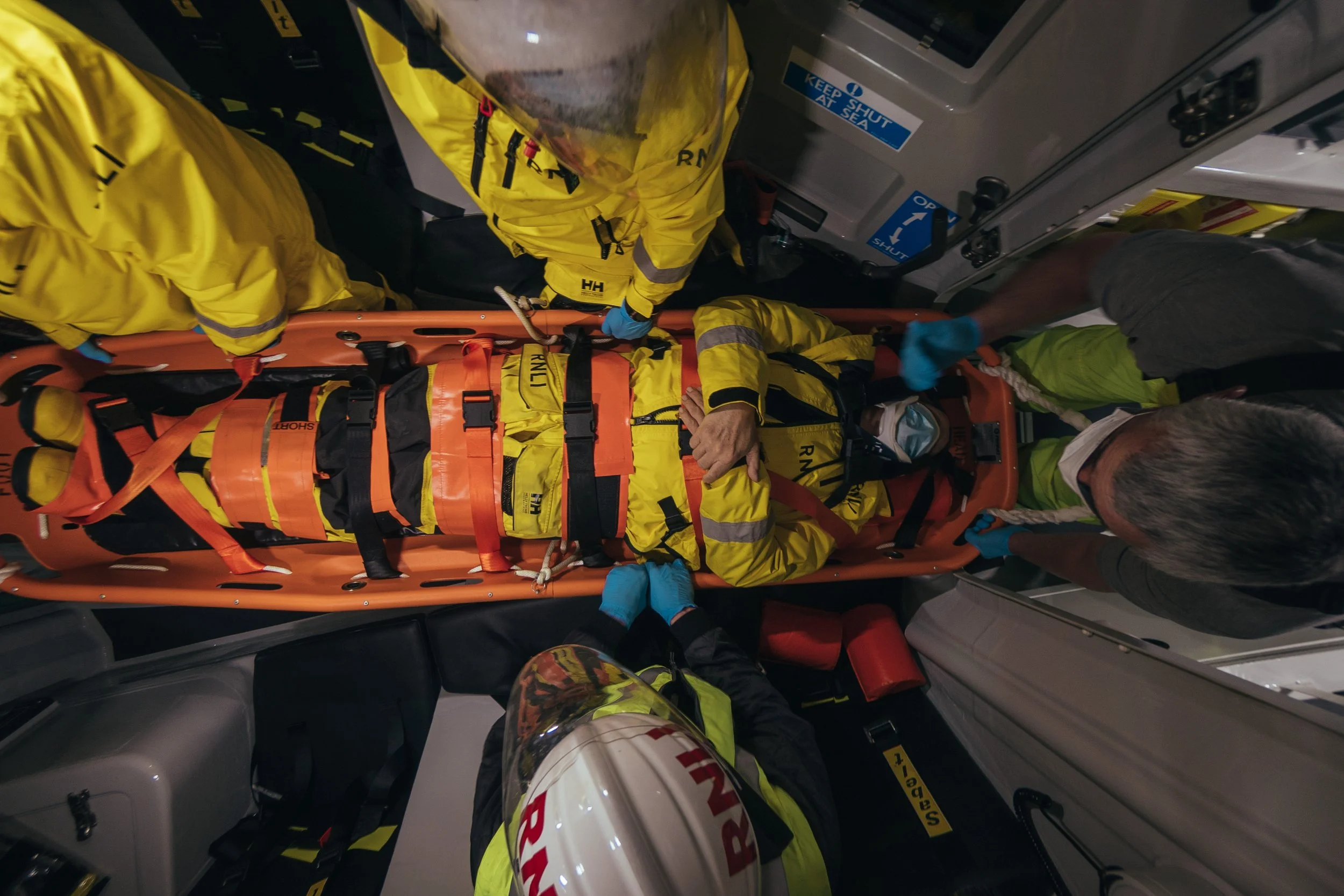 Emergency medical team transporting a patient on a stretcher inside an ambulance, with staff attending to the patient wearing yellow and gray uniforms and blue gloves.