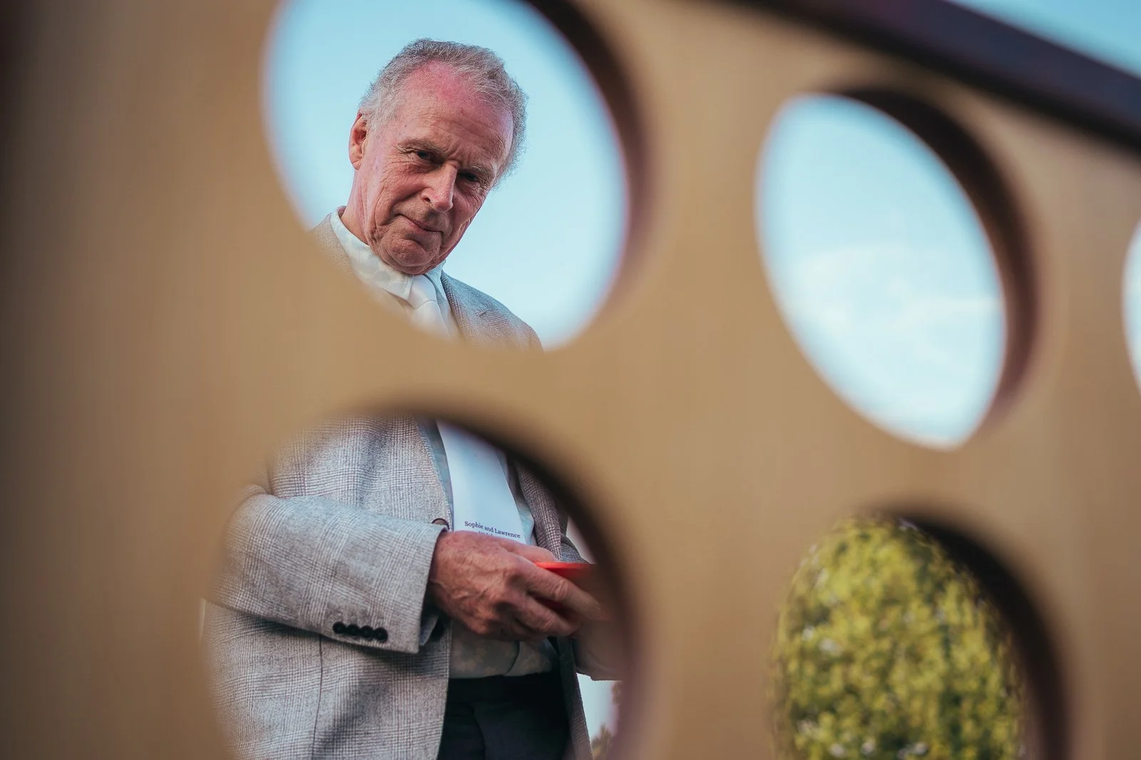 A man with white hair looking through a wooden structure with circular cutouts, viewed from below, holding a red object in his hand, with blue sky and green trees in the background.