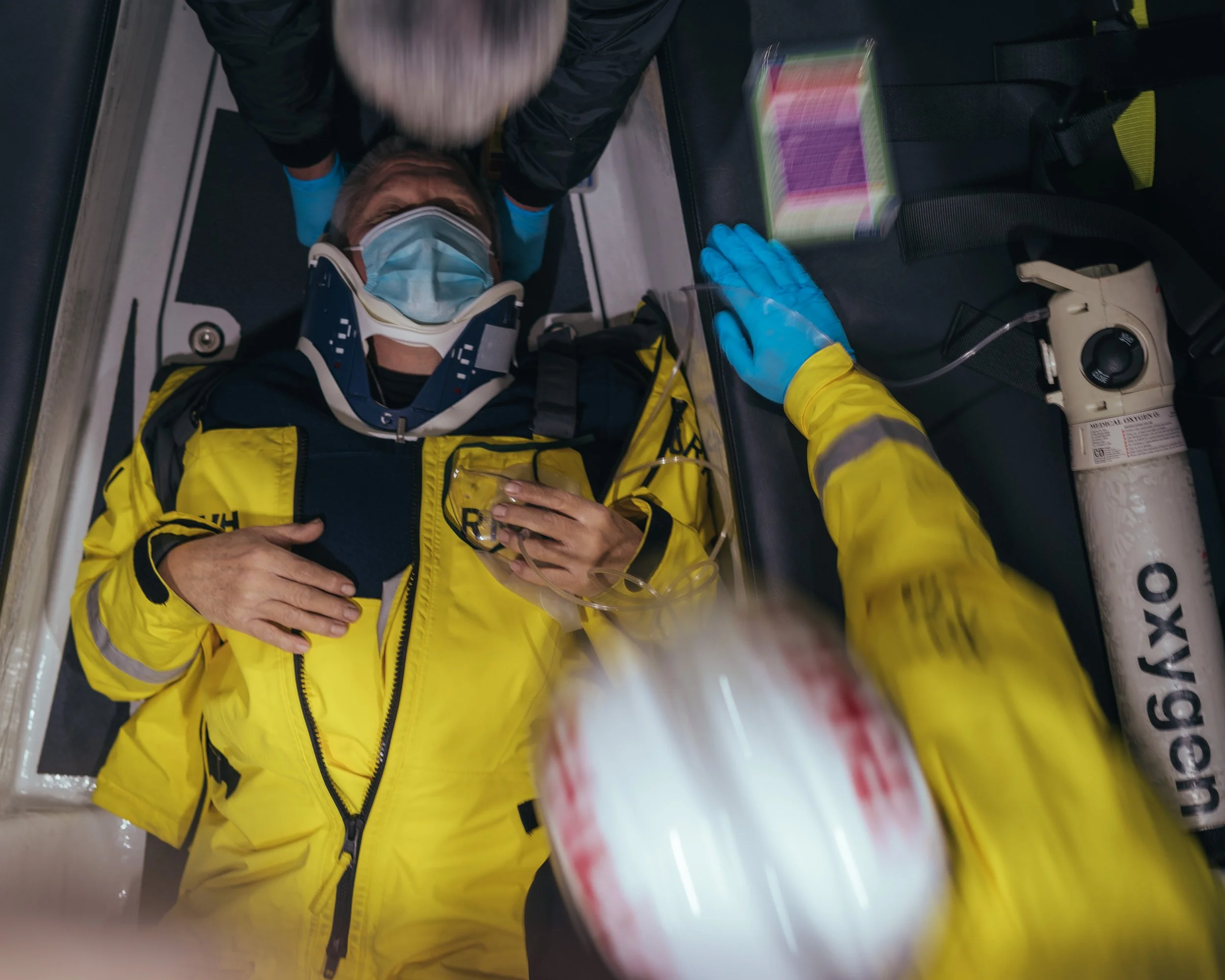 Paramedic in yellow jacket assists injured person lying on stretcher, wearing a cervical collar and mask, in emergency medical setting.