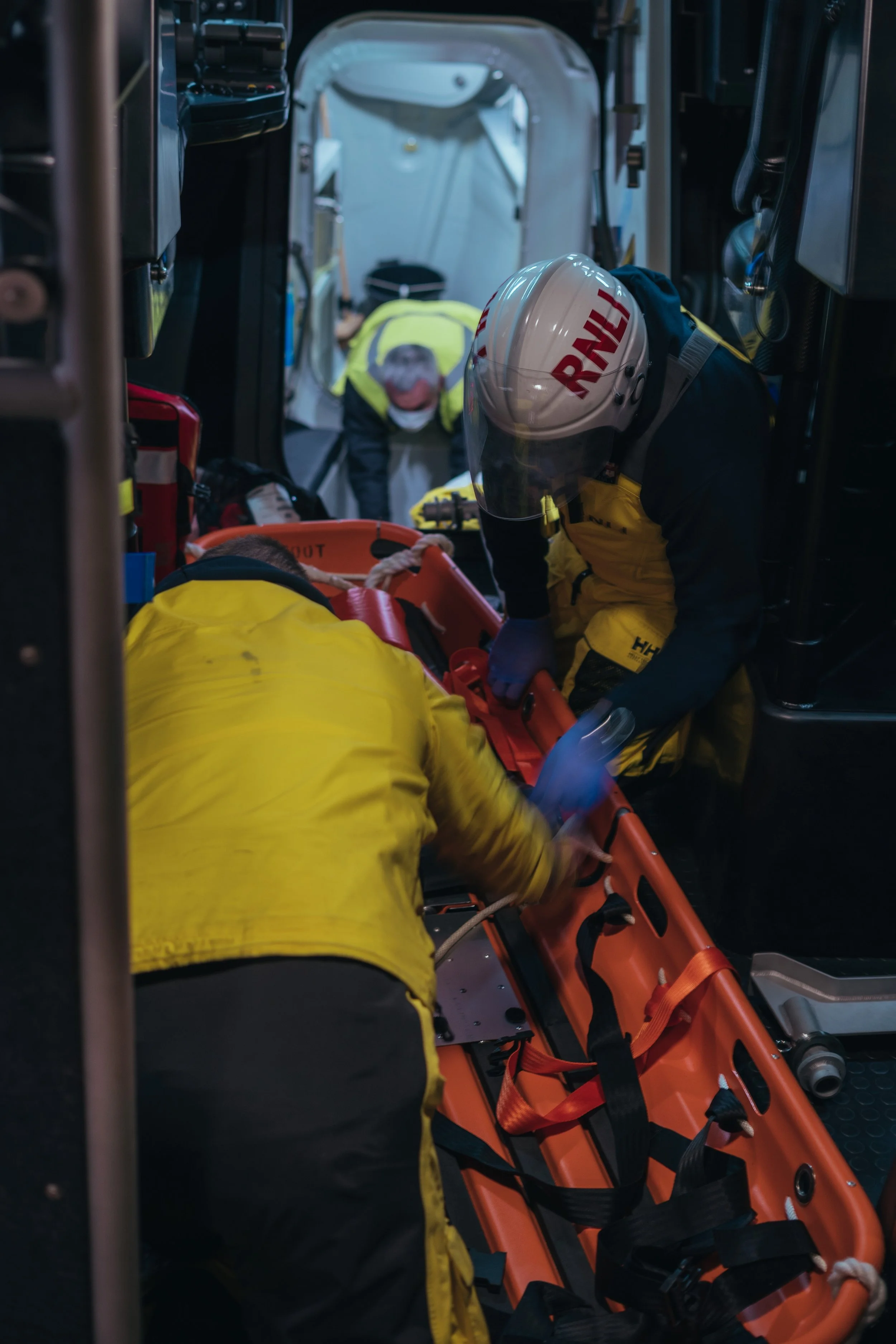 Emergency responders attending to a patient inside an ambulance, with one responder wearing a helmet and yellow jacket, and others assisting on a stretcher.