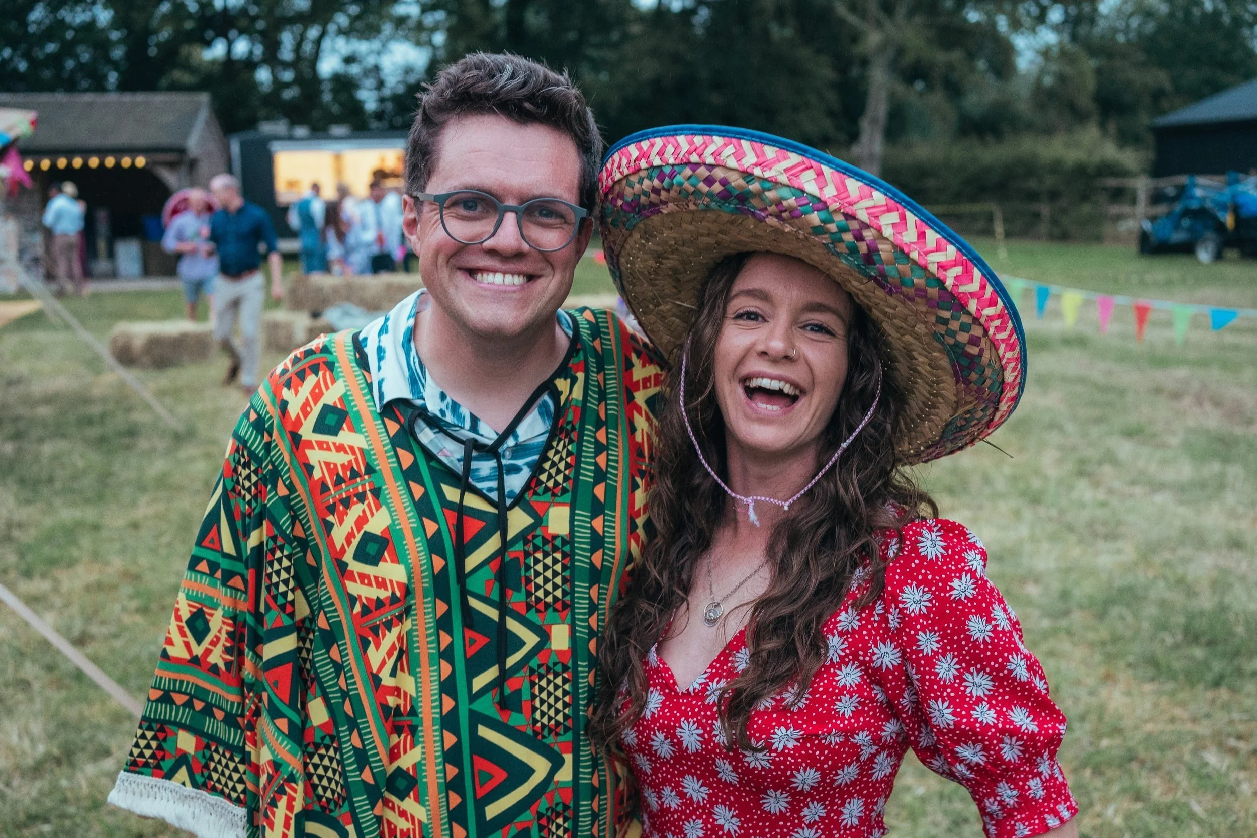 A man and woman smiling at an outdoor festival wearing colorful, casual clothing. The woman has on a large sombrero and a red dress with white floral patterns. The background shows a grassy area with people, hay bales, and bunting flags.
