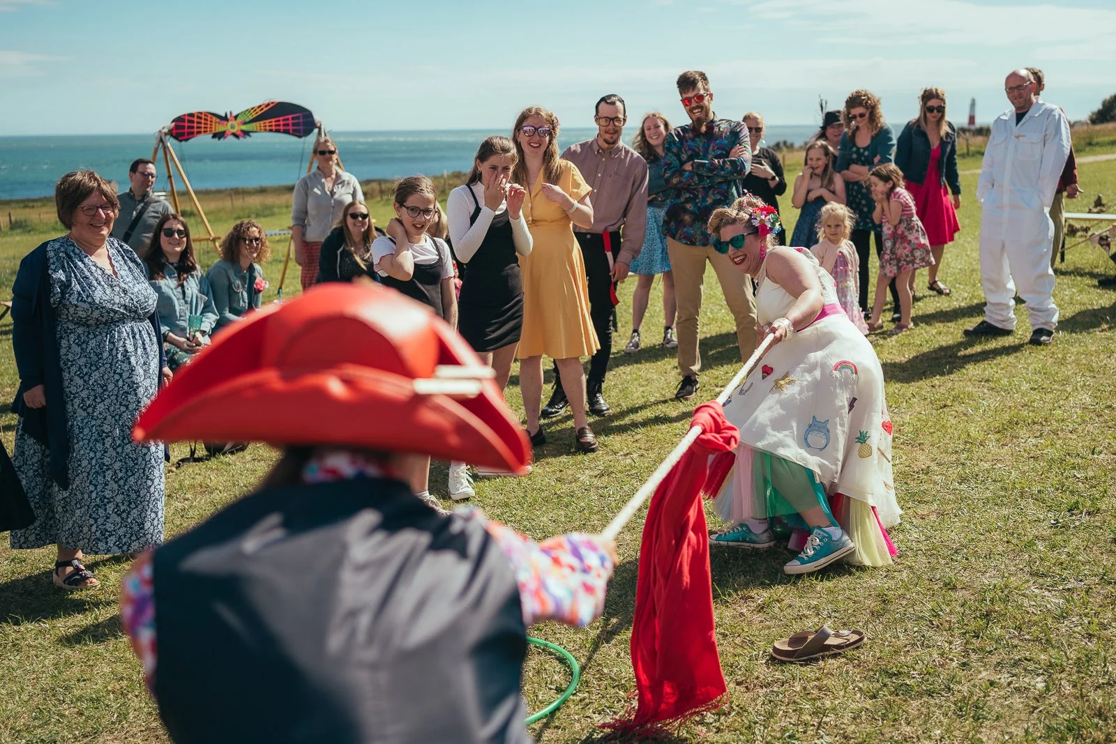People at an outdoor party by the ocean watching a humorous game of tug of war with one person wearing a large red hat and colorful outfit, and another woman dressed in a white dress with embroidered patches.