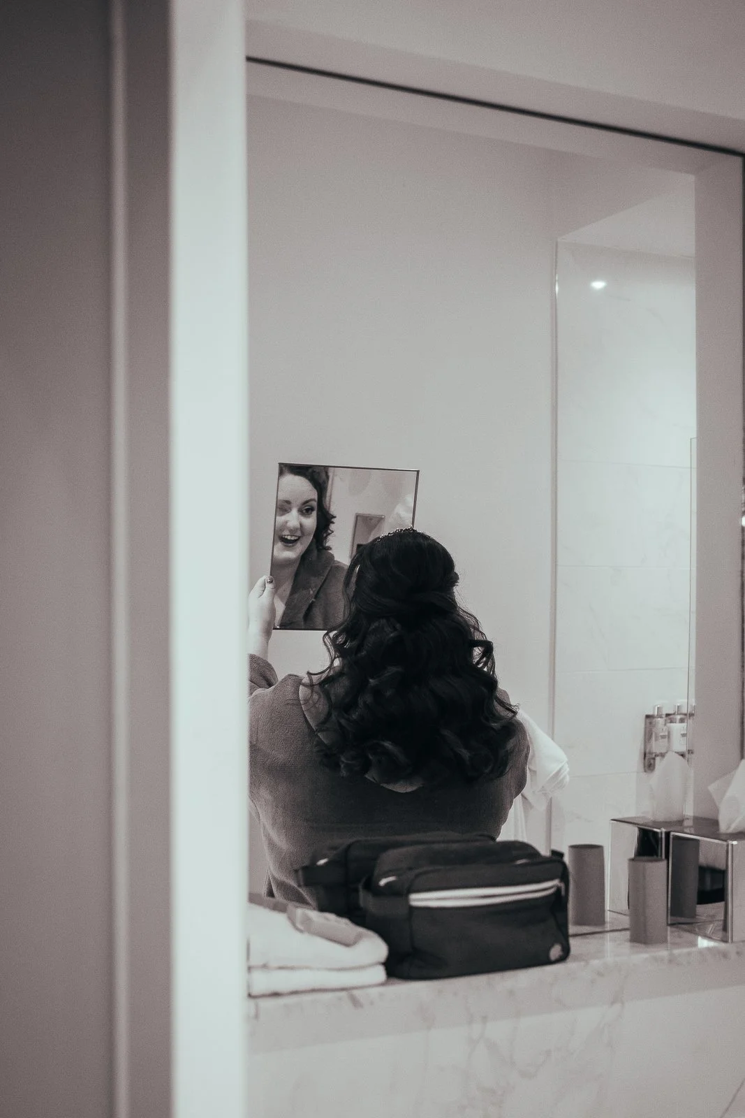 A woman with long dark curly hair looking into a mirror, smiling, with her reflection visible, as she is seated at a bathroom or dressing room vanity with toiletries and a mirror.