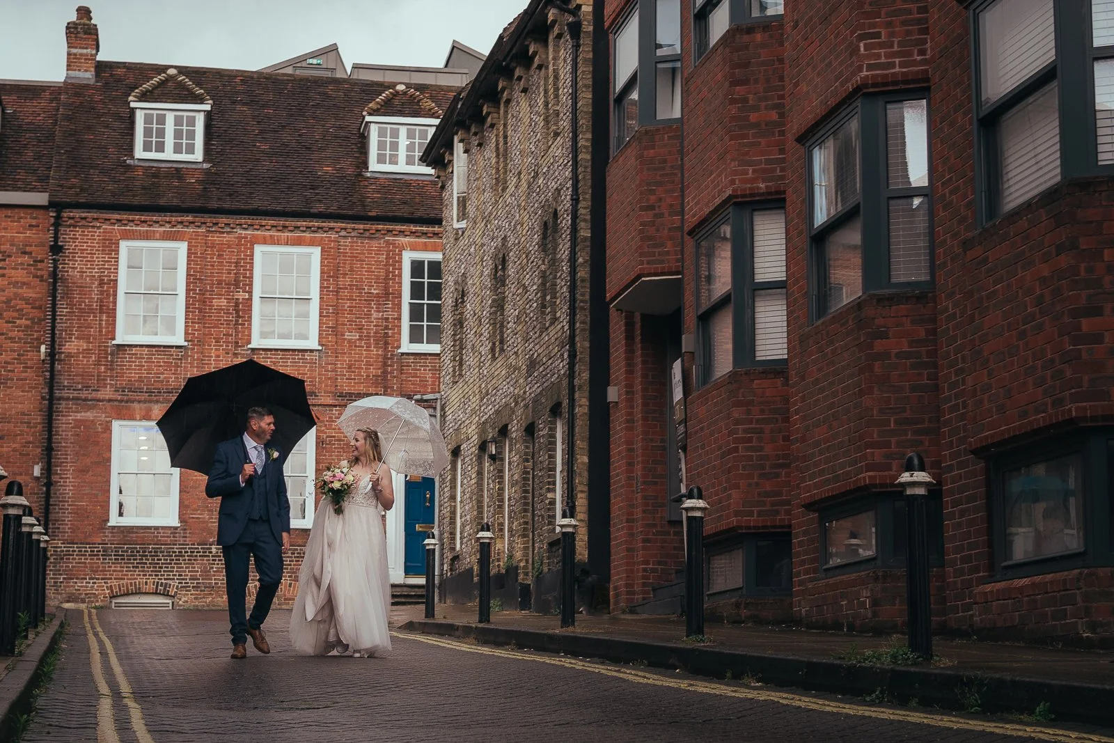 A bride and groom walking together on a rainy street, holding umbrellas. The bride wears a white wedding dress and holds a bouquet, while the groom is dressed in a navy suit. They are engaging in conversation and smiling.