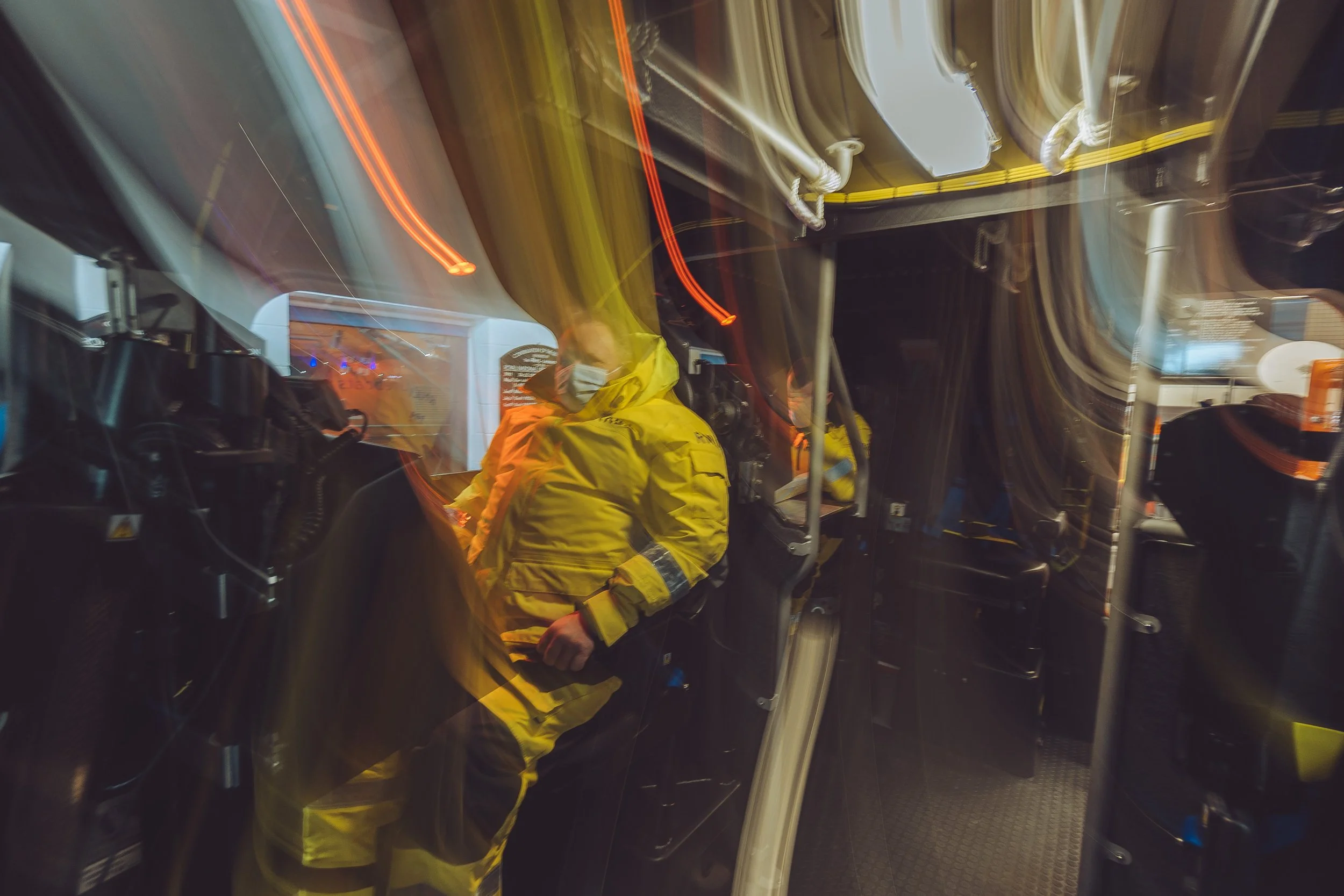 Emergency responder wearing a yellow uniform and face mask inside a fire truck at night, with light trails from moving lights creating a blurred effect.