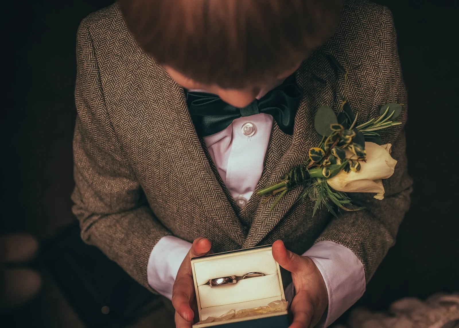 A man dressed in a herringbone blazer with a white shirt, black bow tie, holding an open ring box with two wedding bands and a boutonniere attached to his blazer. The photo is taken from above.