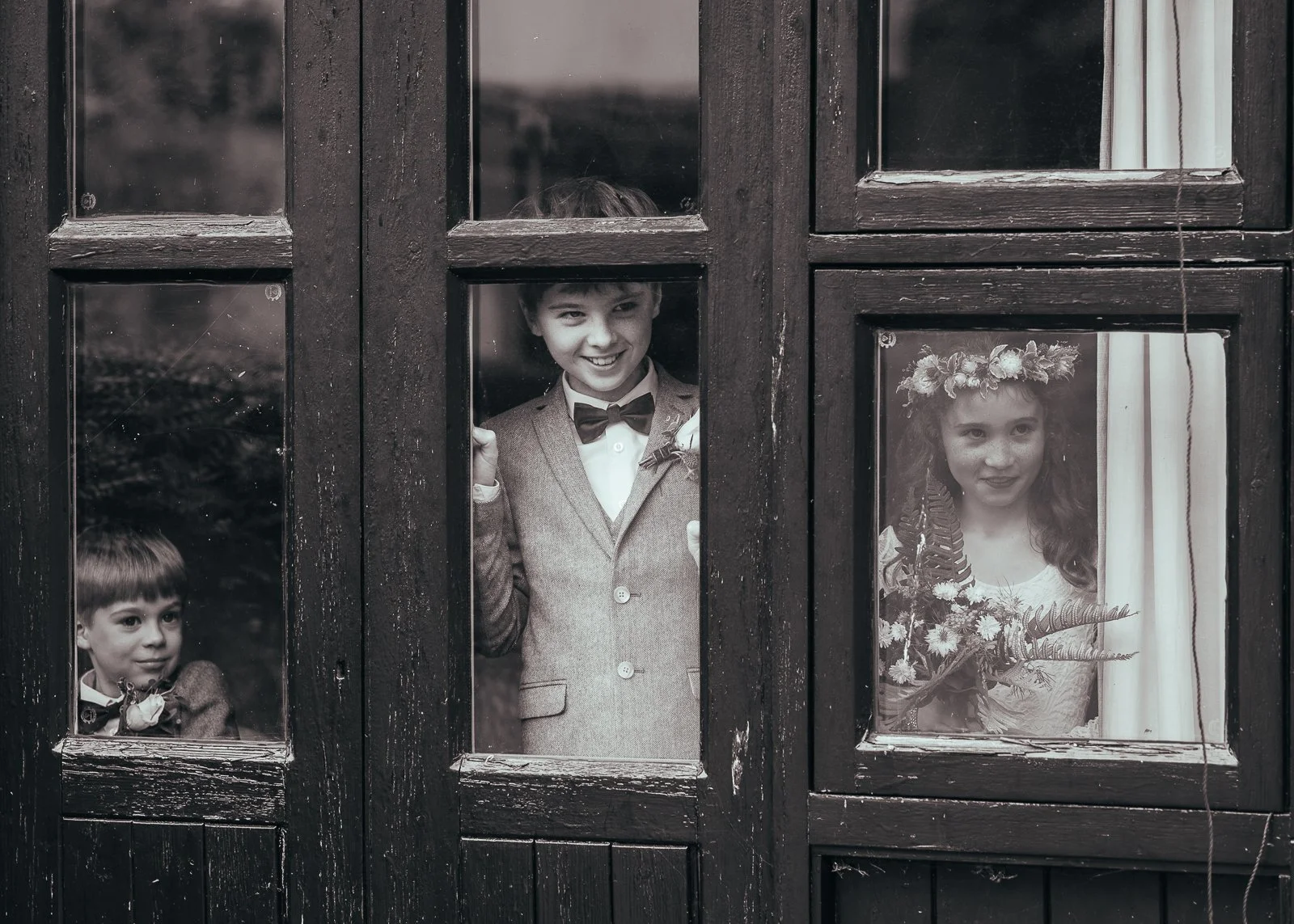 Three children peering through a window, two boys and one girl, dressed in formal attire, with the girl wearing a flower crown, during a special occasion.