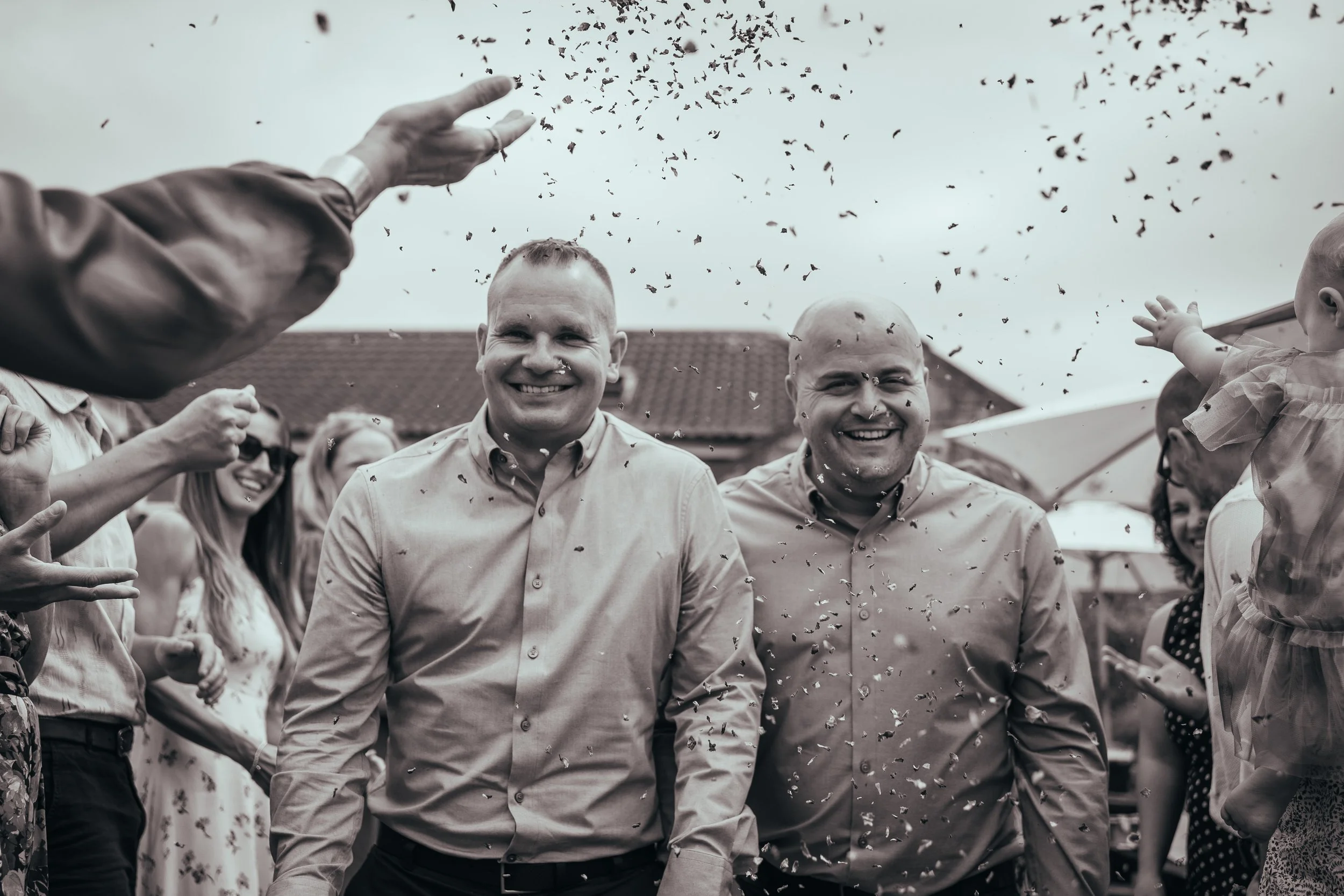 Two grooms being covered with confetti. Black and white candid wedding image.