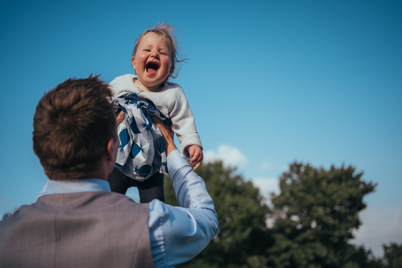 A man holding up a laughing young girl outdoors with a blue sky and trees in the background.