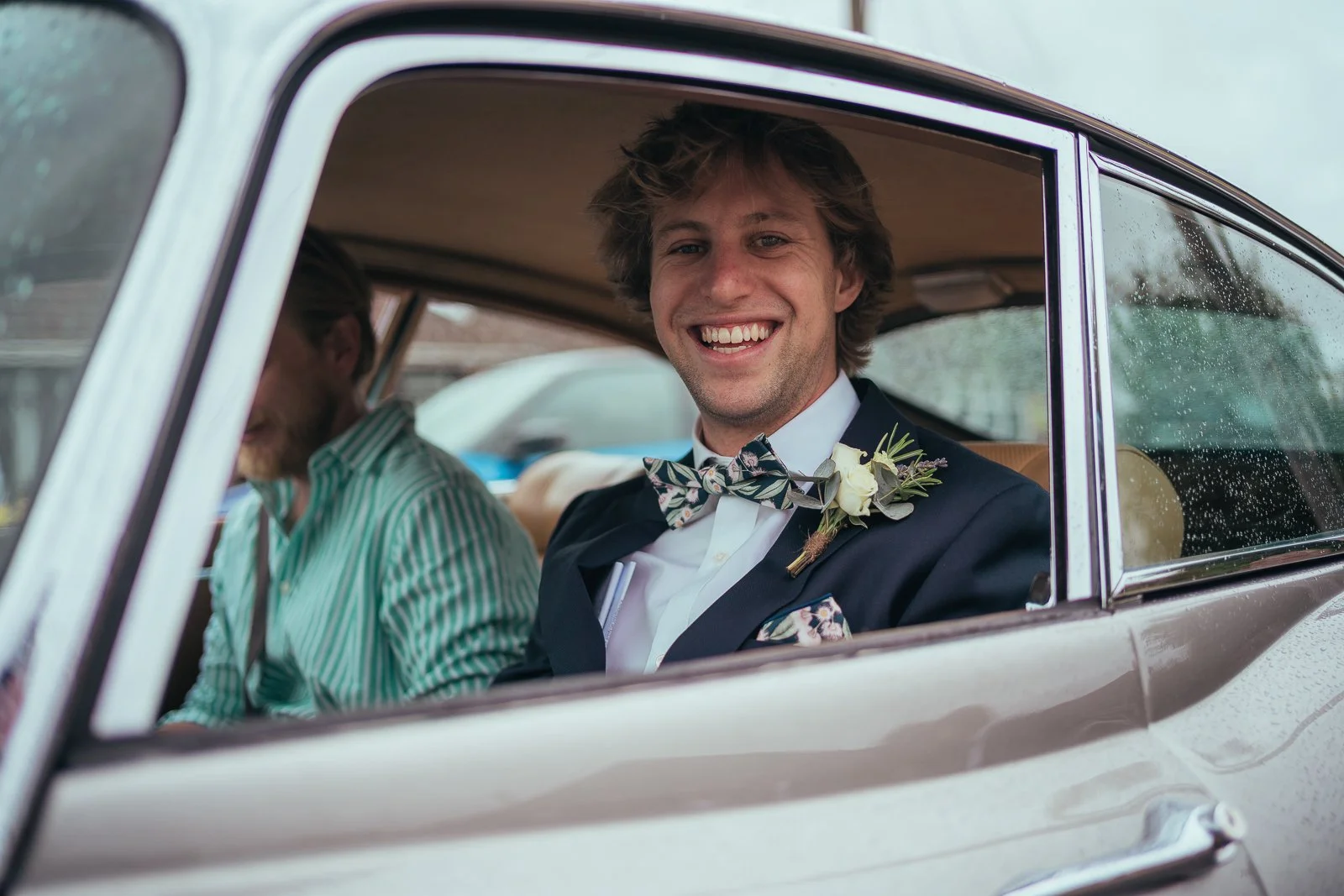 Man in suit with floral bow tie and boutonniere smiling while sitting in vintage car
