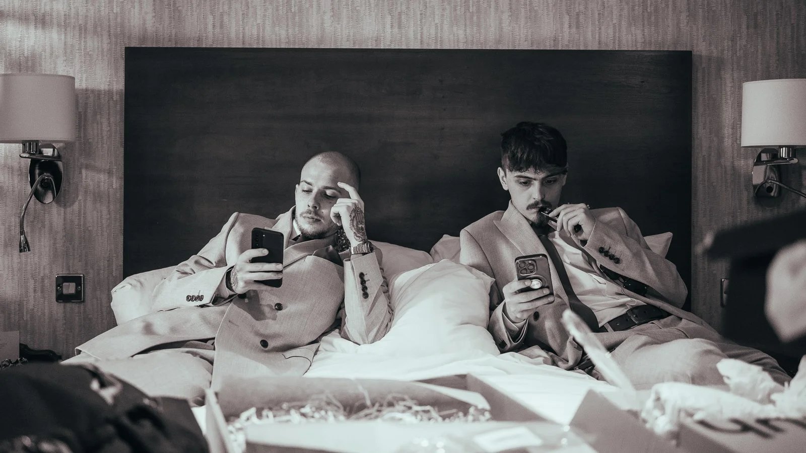 Two men in suits sitting on a hotel bed, both looking at their smartphones, with room lamps on either side of the headboard.