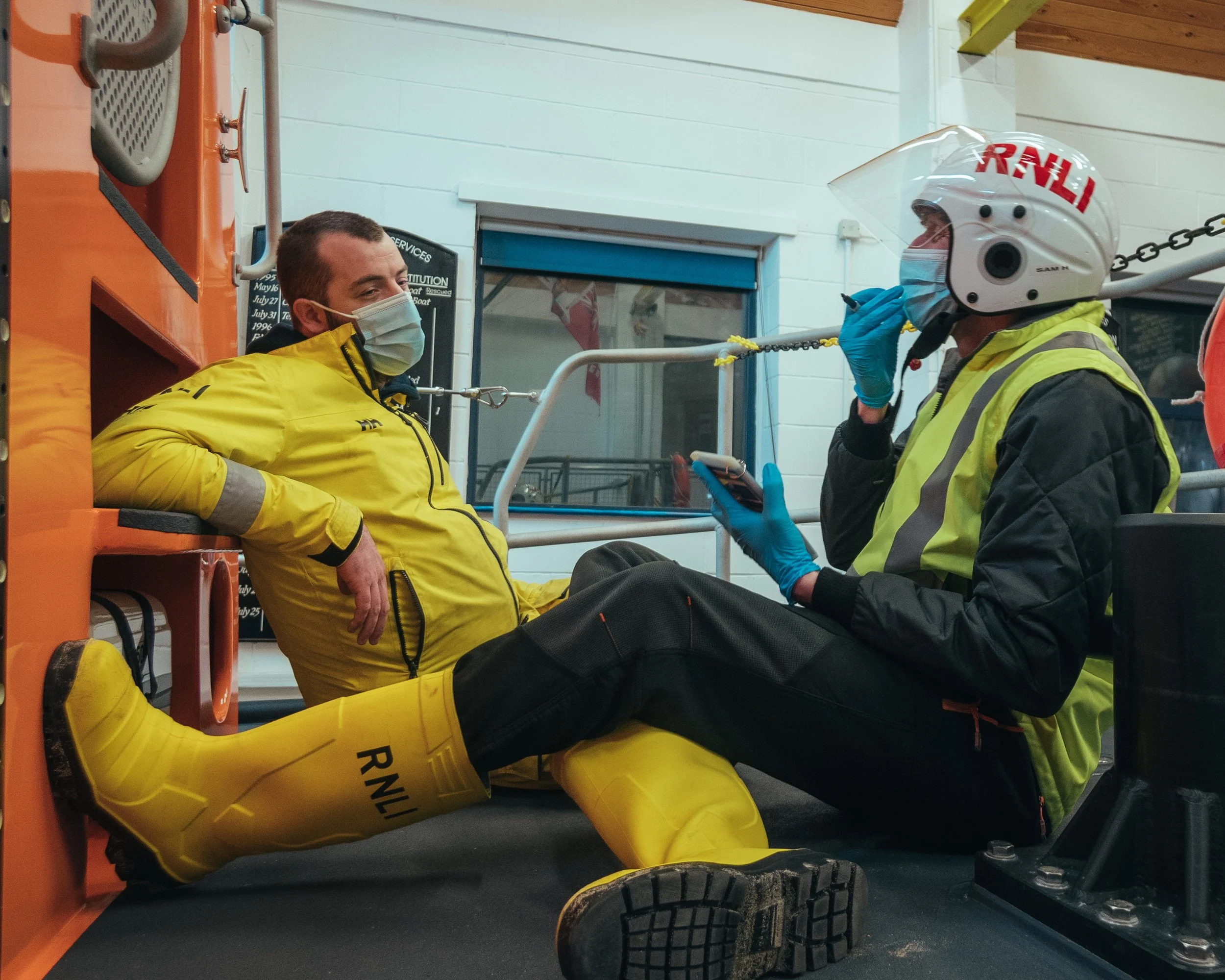Two men wearing masks, one in a yellow rain jacket and rain boots, sitting on the ground in a room with safety equipment, talking. The man on the right has a helmet with 'RNLI' written on it, a hi-vis vest, and blue gloves, and is holding a smartphon