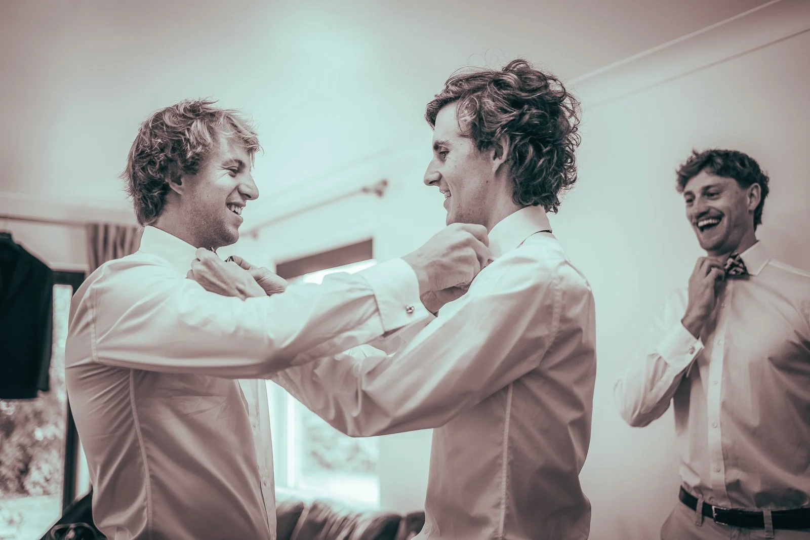 Three men in formal shirts getting ready, with two of them adjusting their ties and one smiling at the process.