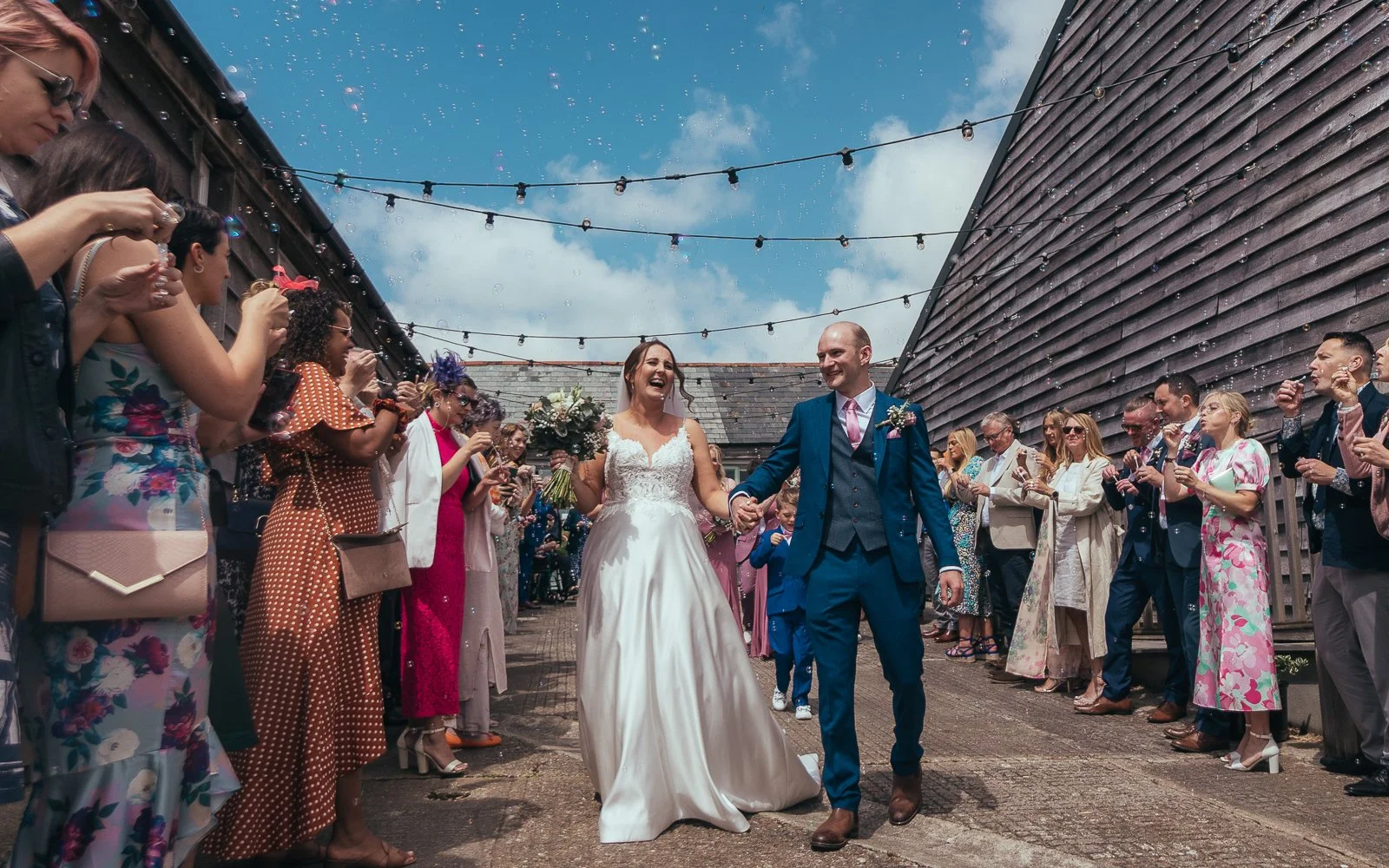 Bride and groom walking hand in hand, smiling, with wedding guests in colorful attire on either side, under string lights on a bright day.