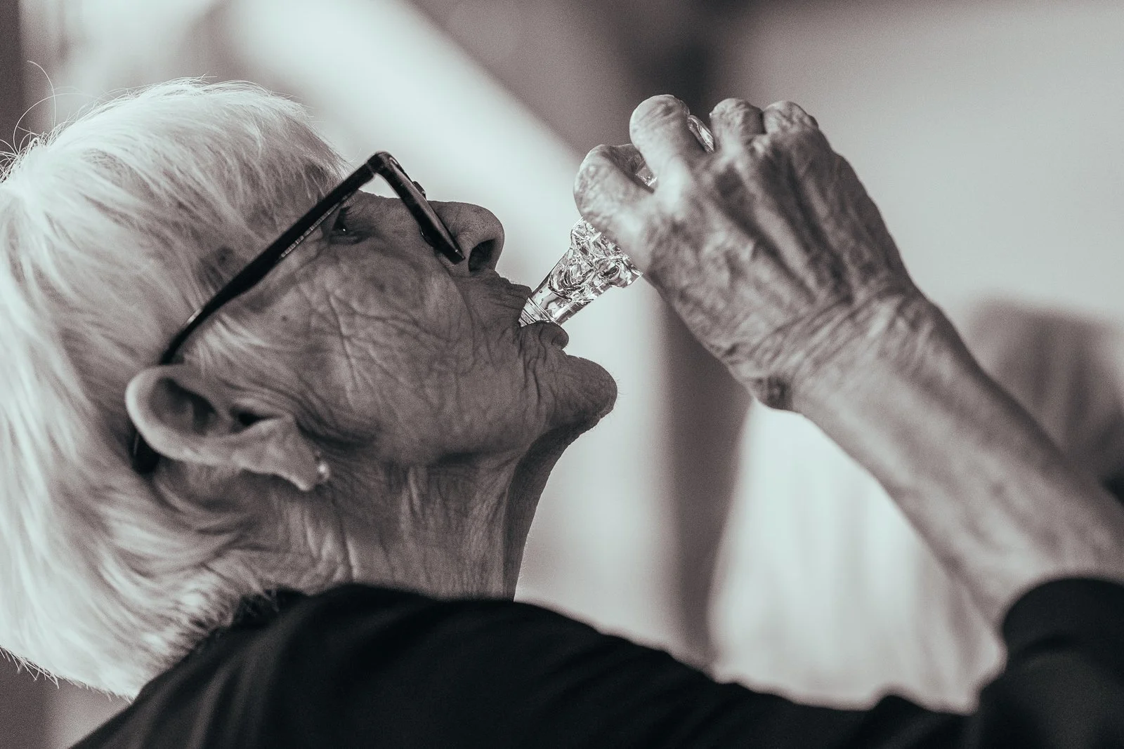 An elderly woman with short white hair and glasses drinking gin from a minature bottle given as a wedding favour with her head tilted back.