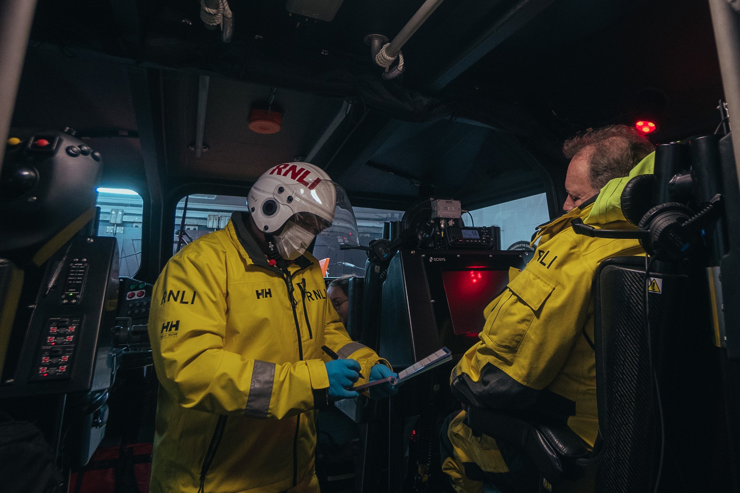 Two emergency responders inside a vehicle, wearing yellow jackets with 'RNLI' logos, one reading a notepad and the other sitting with eyes closed, with equipment surrounding them.