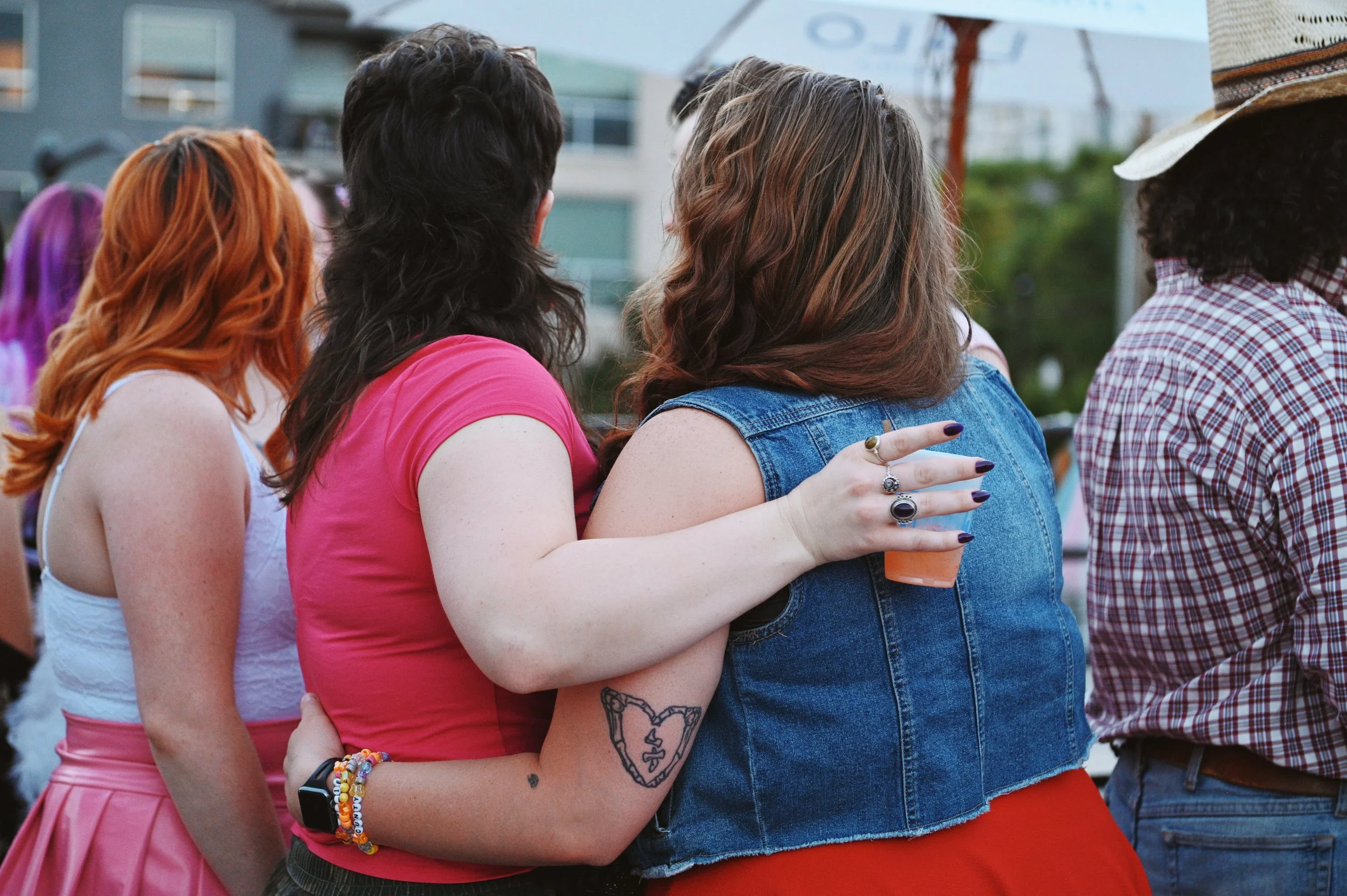 A group of people at an outdoor event, with two women embracing. One woman has red hair and is wearing a white top, while the other has brown hair with highlights and is wearing a denim vest, holding a drink.