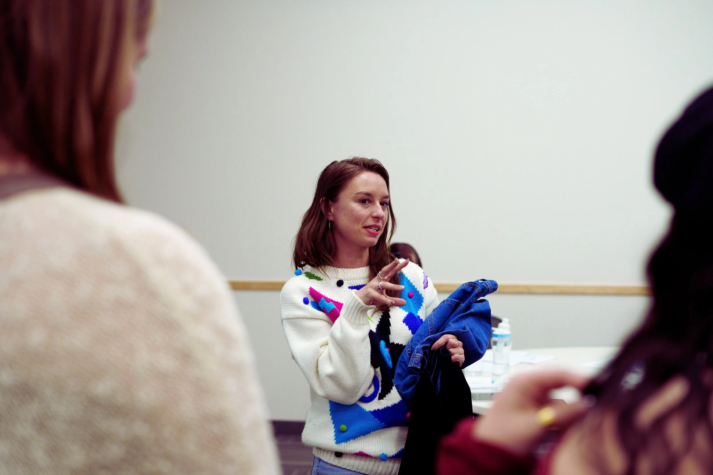 A woman with reddish-brown hair wearing a options colorful patterned sweater, engaged in conversation in a meeting room with a plain white wall in the background.