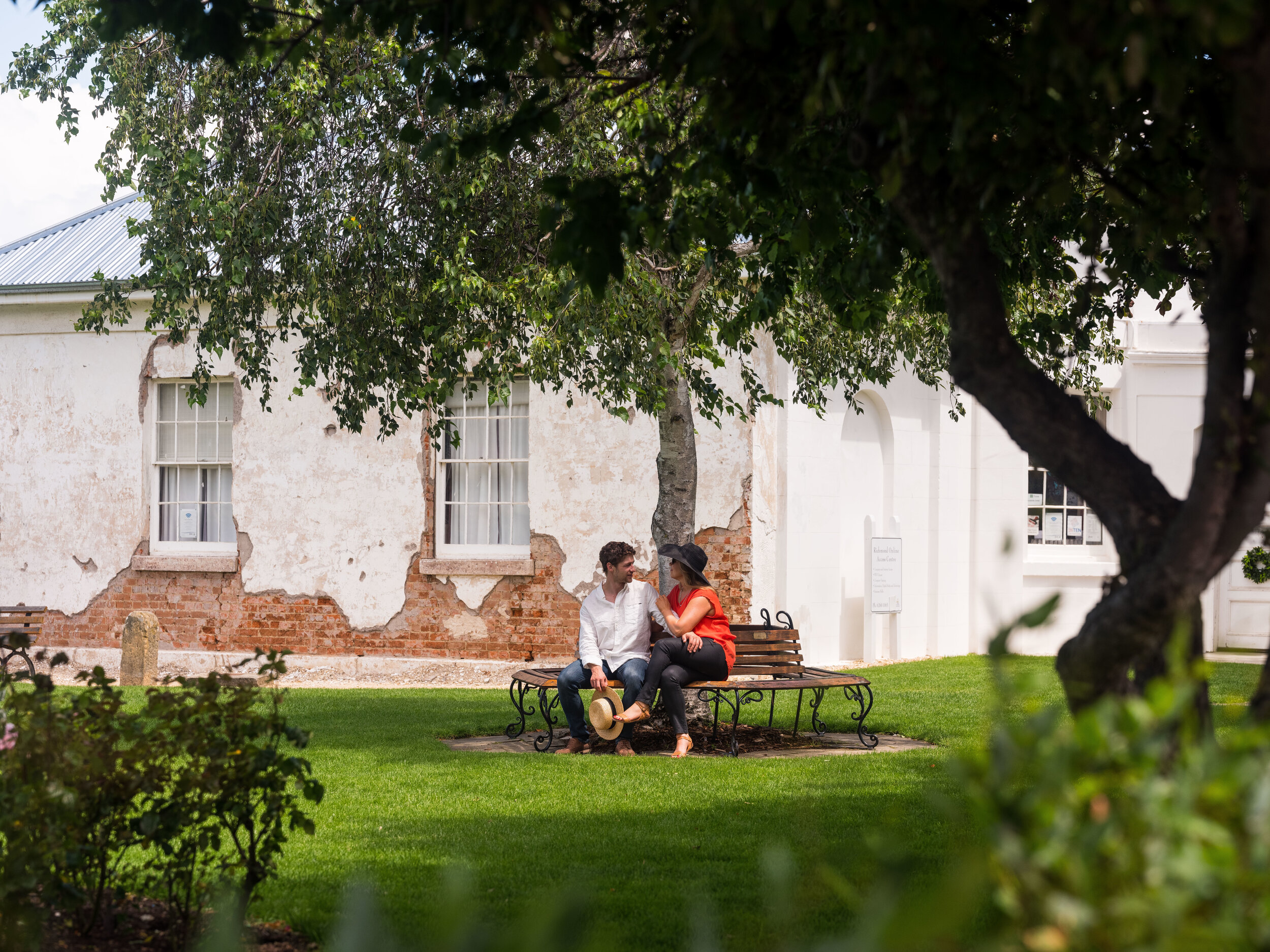 A couple sitting on a park bench in front of an old building with peeling paint. They are surrounded by greenery and trees.