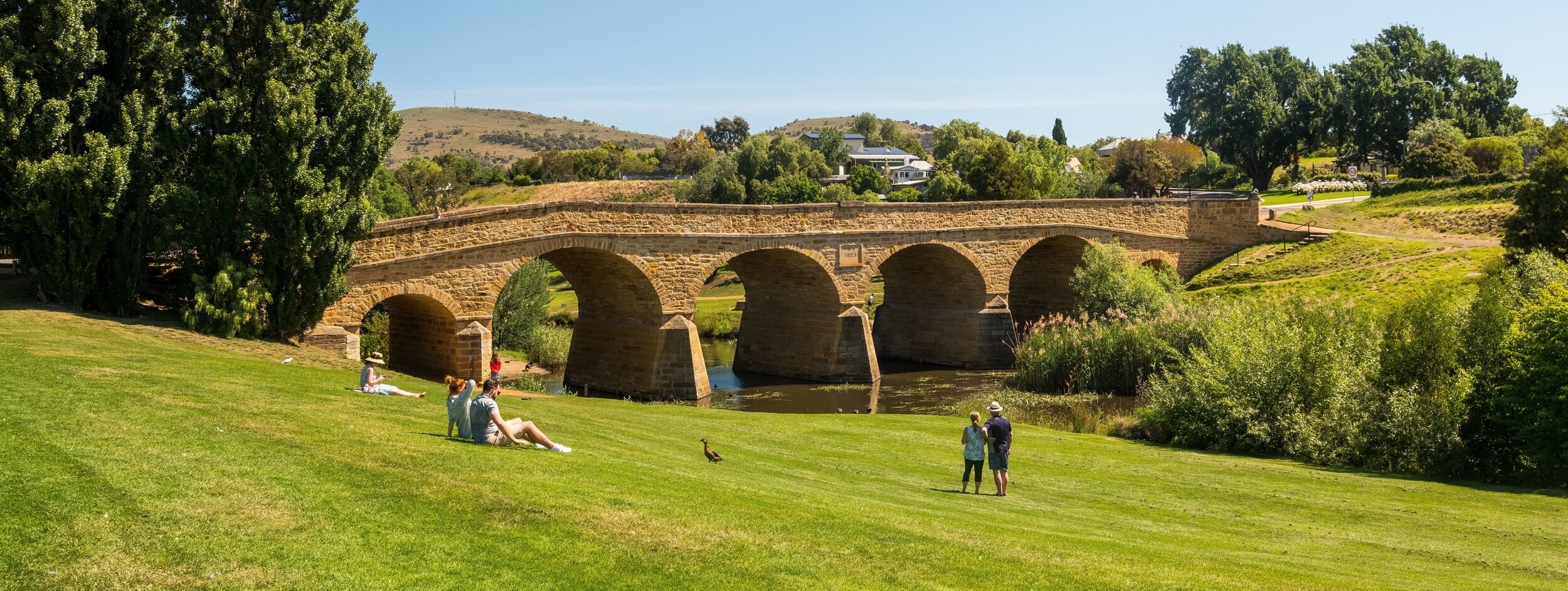 Historic stone bridge with arches spanning over a river, surrounded by green grass and trees, with people relaxing nearby.
