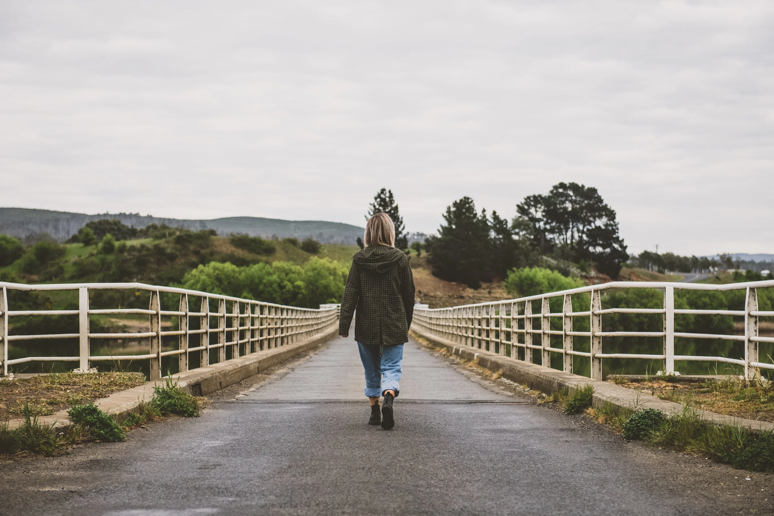 Person walking on a bridge surrounded by greenery under a cloudy sky
