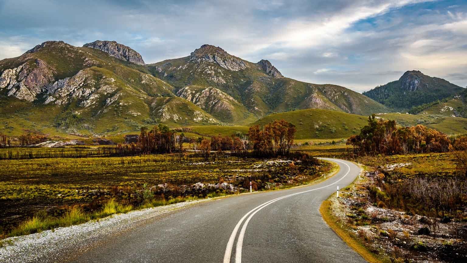 Winding road leading towards jagged, green mountains under a partially cloudy sky.