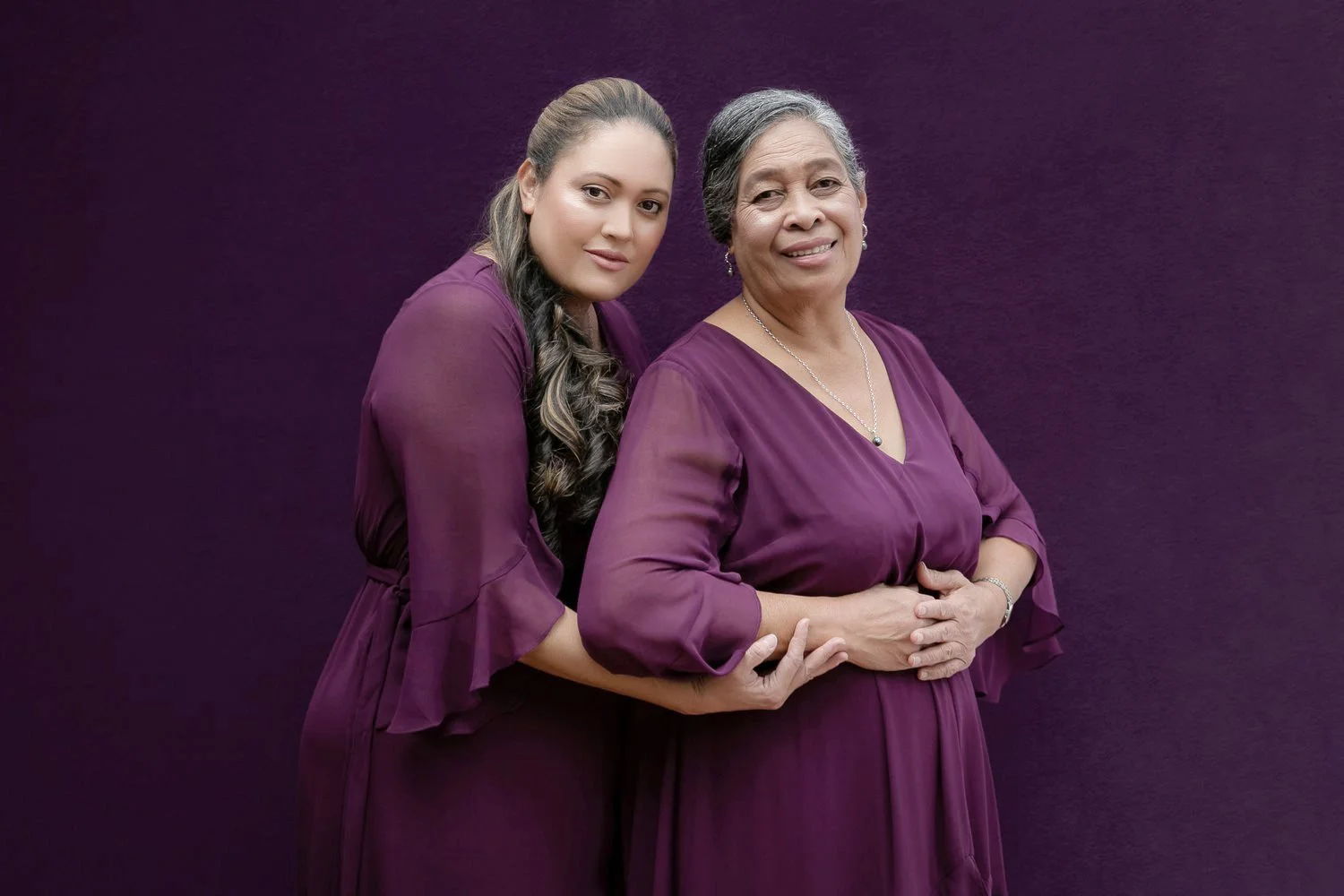Two women standing against a purple background, both wearing purple dresses. One woman with long hair embraces the other, who has short hair.