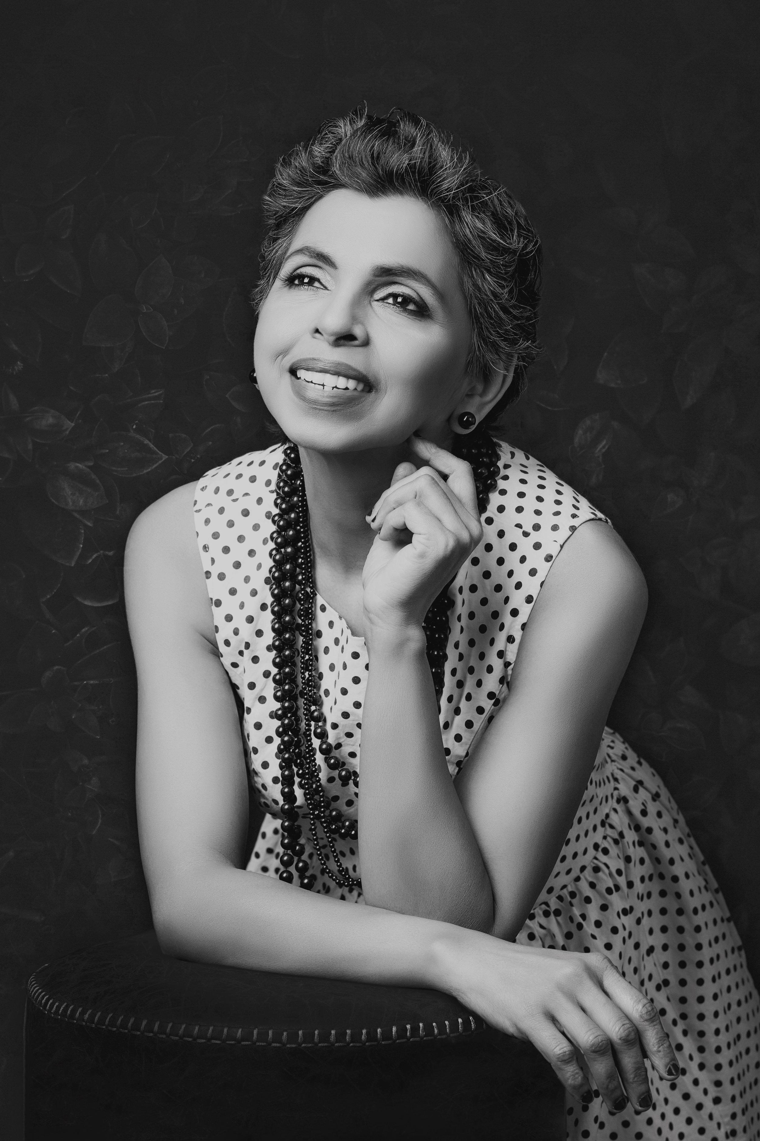 Black and white photo of a smiling woman in a polka dot dress, resting her chin on her hand.