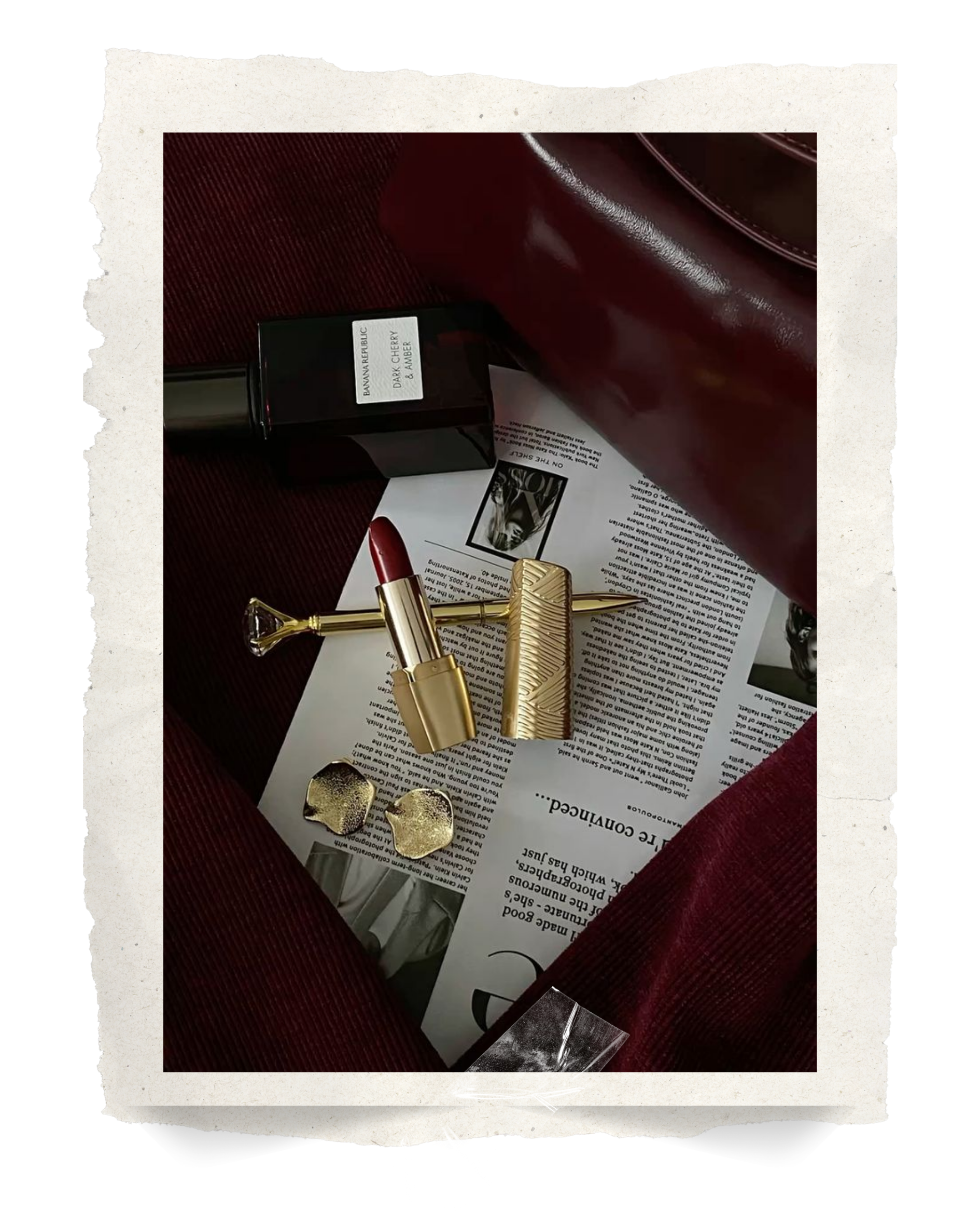 Close-up of various makeup and fashion items on a red surface, including a lipstick, gold earrings, a gold lipstick case, a black bottle labeled 'Dark Cherry & Amber,' and some printed materials.