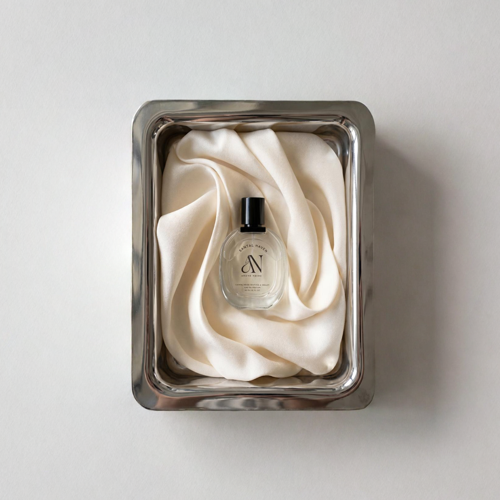 Clear perfume bottle labeled 'Santal Haven' placed on silk fabric inside a silver tray.