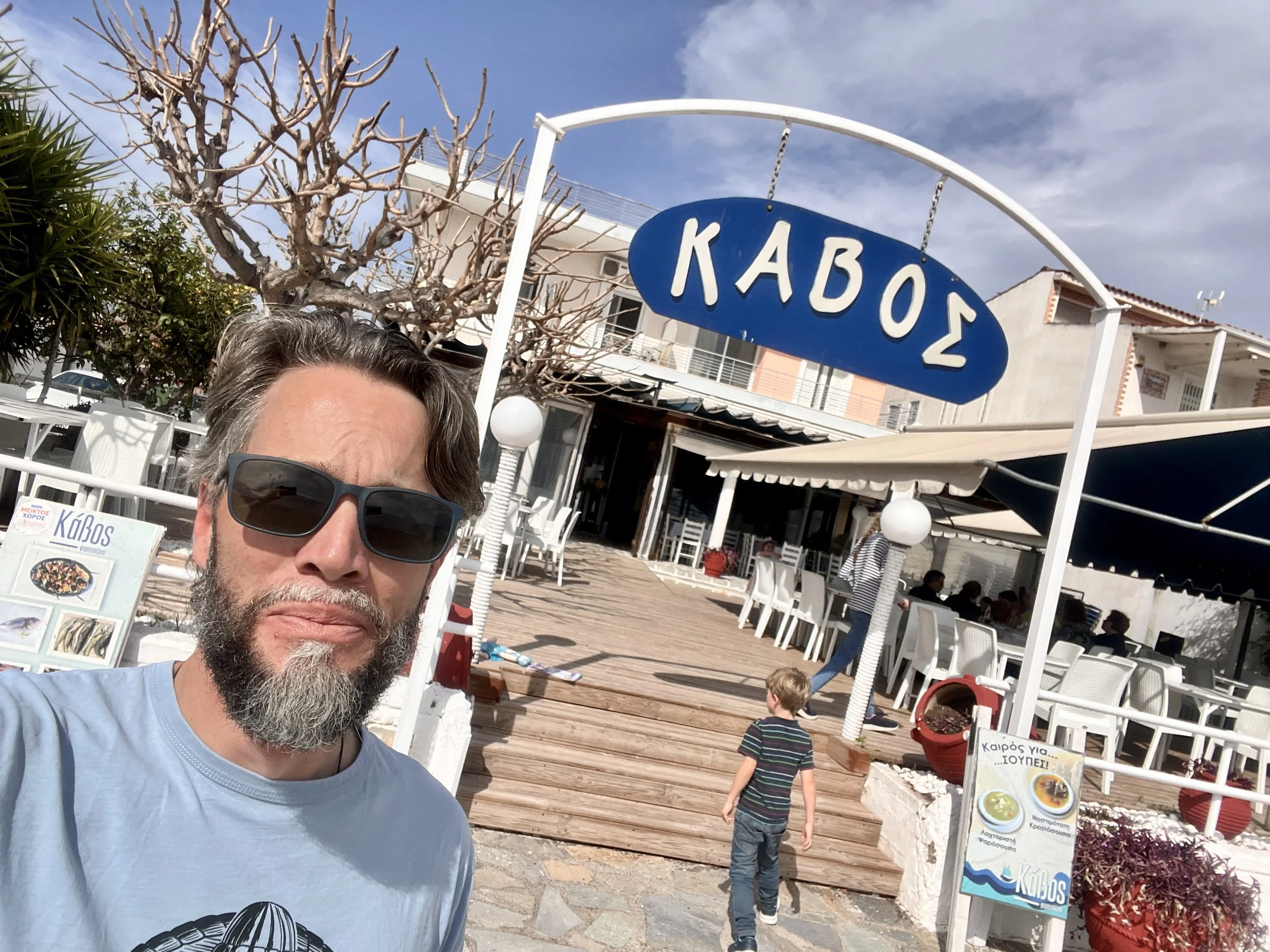 Ein Mann mit Sonnenbrille steht vor einem Restaurant mit dem griechischen Schriftzug "ΚΑΒΟΣ" auf einem Schild. Im Hintergrund sitzt eine Gruppe von Menschen auf einer Terrasse unter einer Markise. Ein Kind läuft in Richtung des Eingangs. Der Himmel i