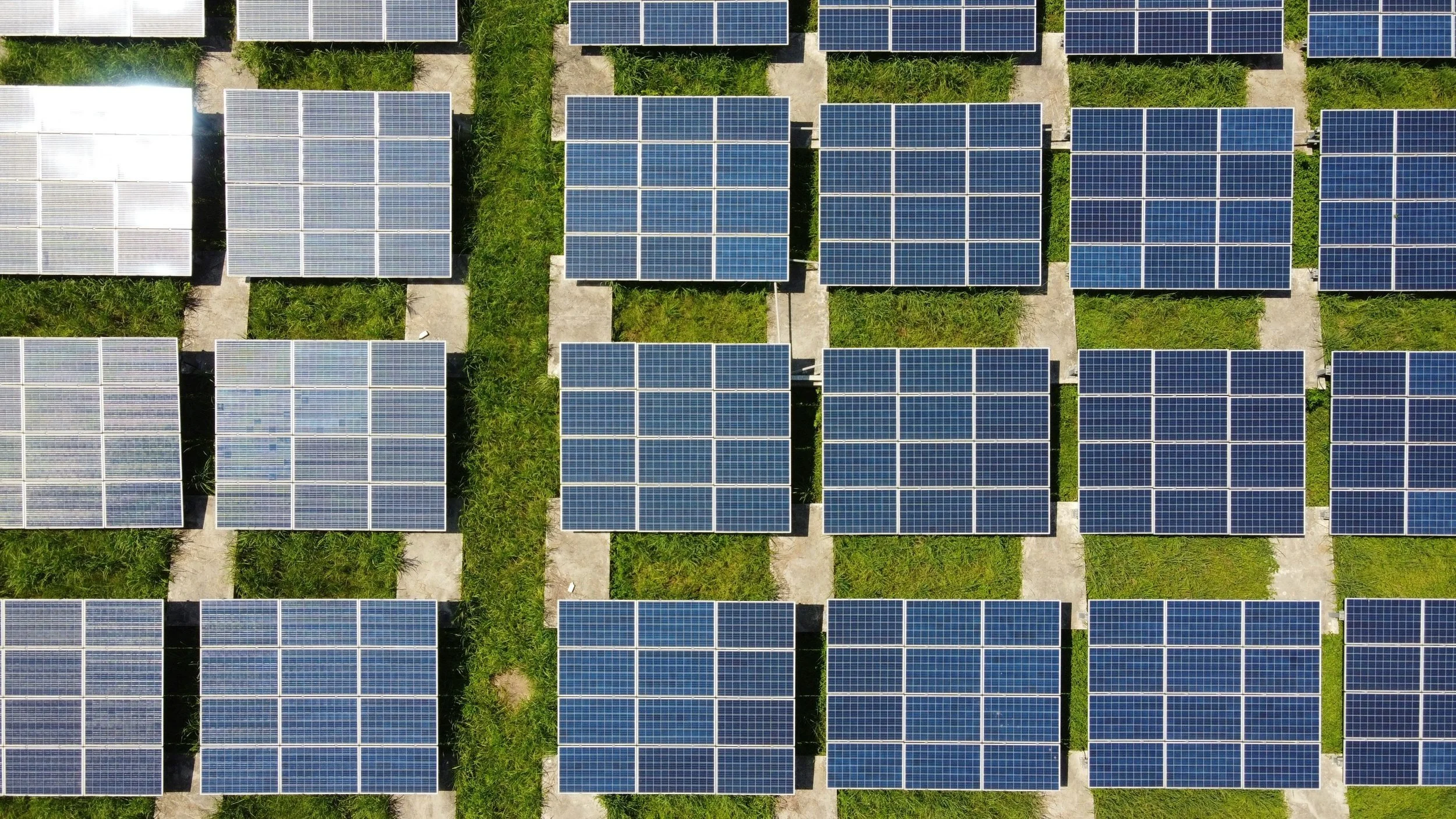 An aerial view of multiple solar panels arranged in rows on grassy plots with pathways between them.