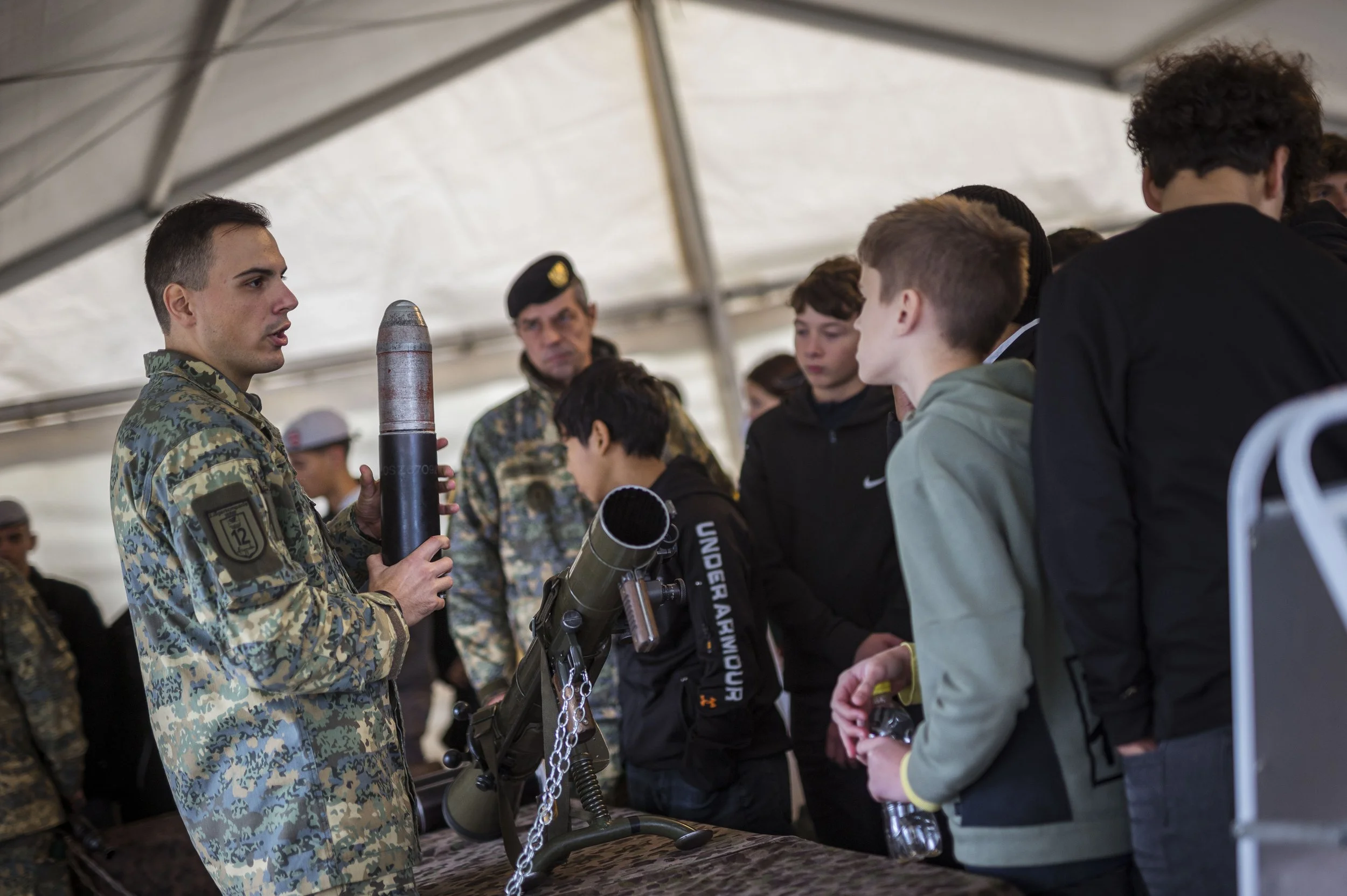 Jugendliche im Gespräch mit einem Soldaten, der eine Handfeuerwaffe und Munition zeigt, in einem Zelt bei einer militärischen Demonstration oder Ausbildung.